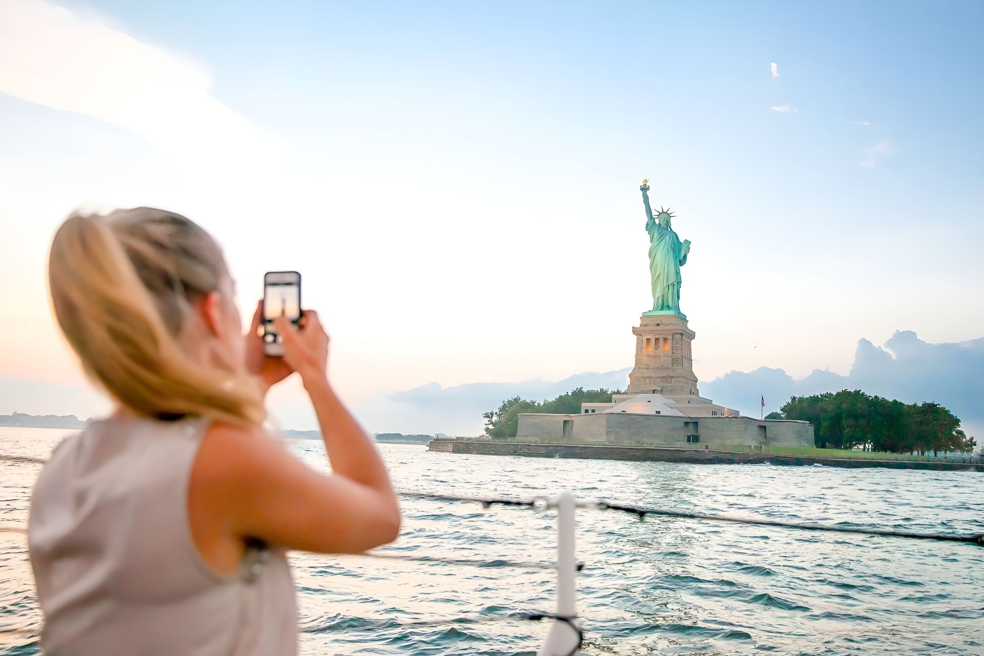 Woman taking a photo of the Statue of Liberty on a sunny day from a boat.