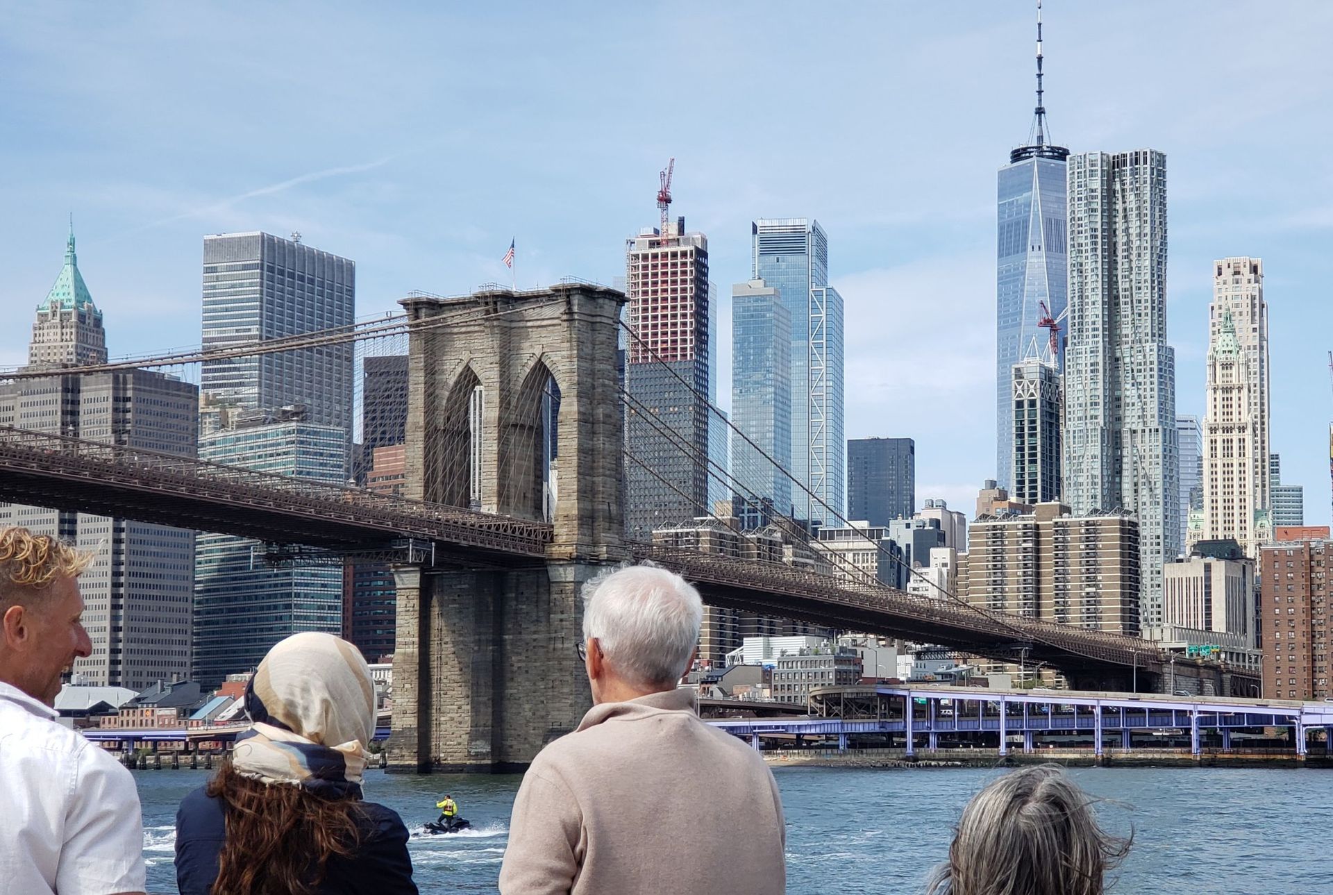 People view the Brooklyn Bridge and NYC skyline from a boat on a sunny day.