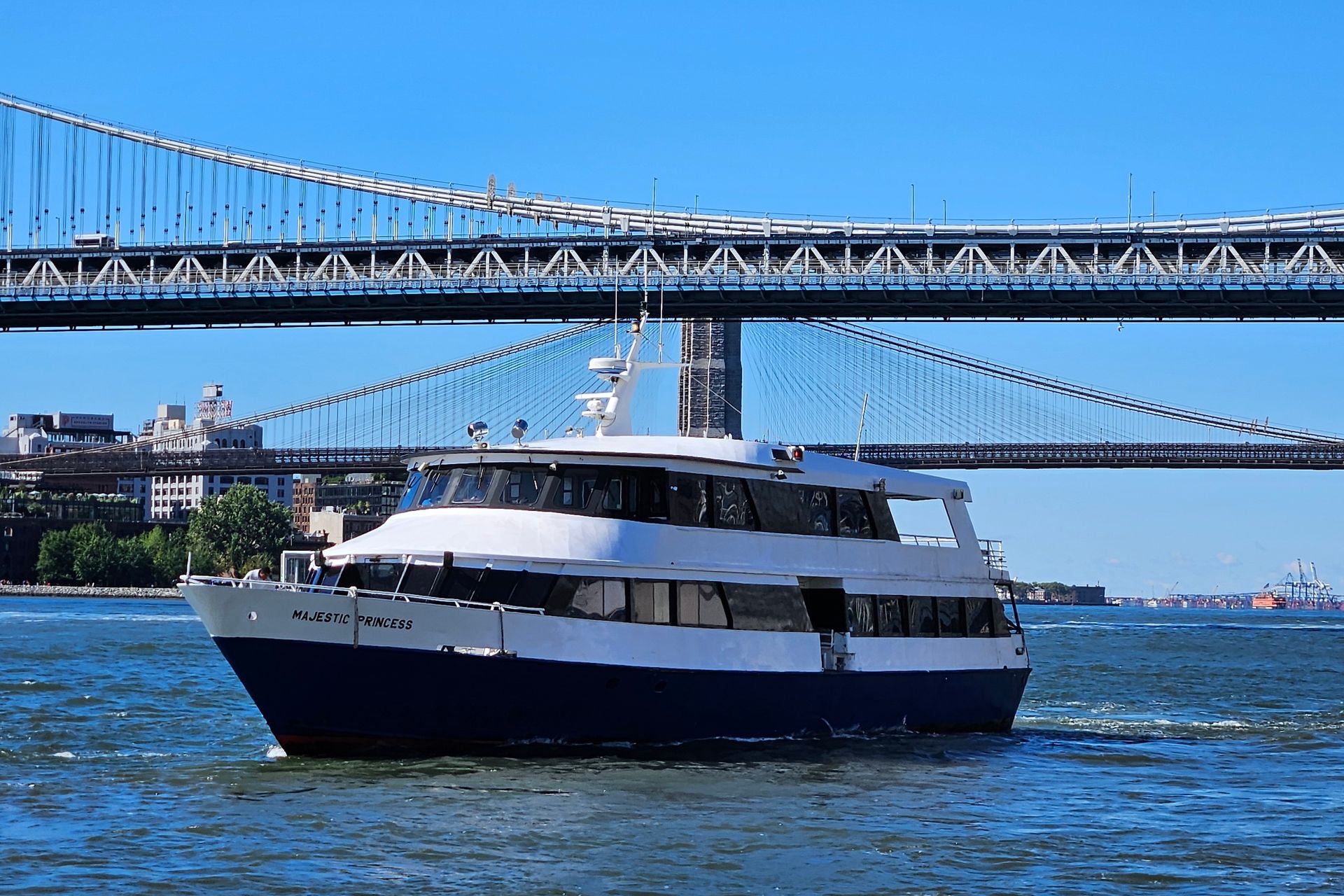 Boat sailing under a bridge on the water, with a blue sky and cityscape in the background.