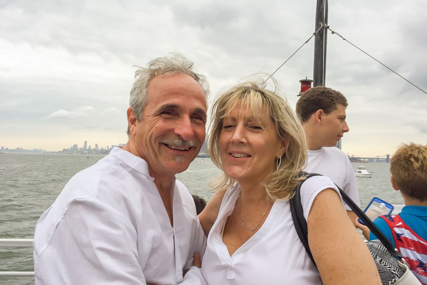 Smiling couple on a boat, near a city skyline. Man with mustache, woman with blond hair; overcast sky.