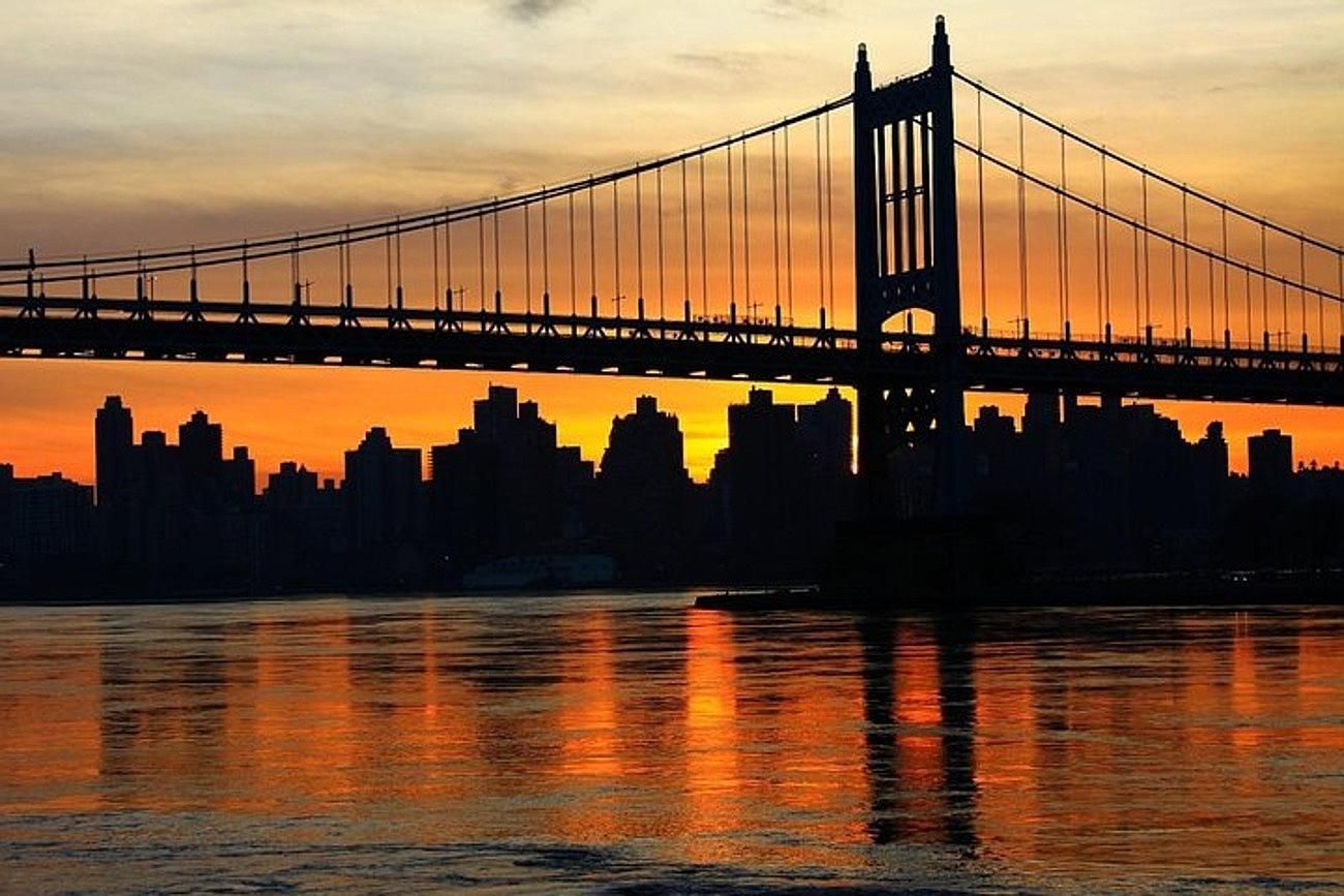 Silhouette of a bridge and city skyline at sunset reflected in the water, orange sky.
