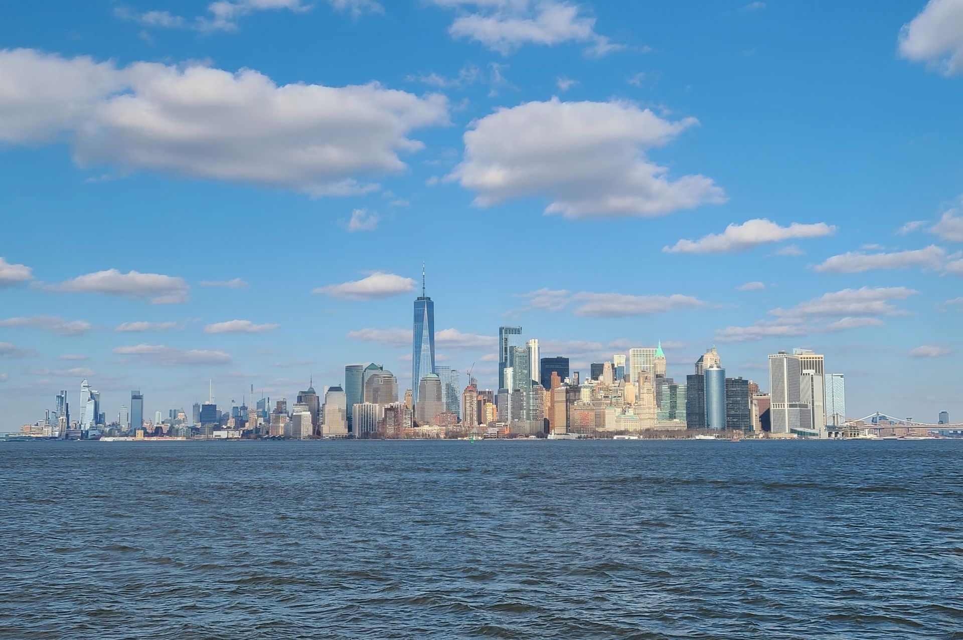 New York City skyline on a sunny day with blue sky and clouds, viewed from across water.