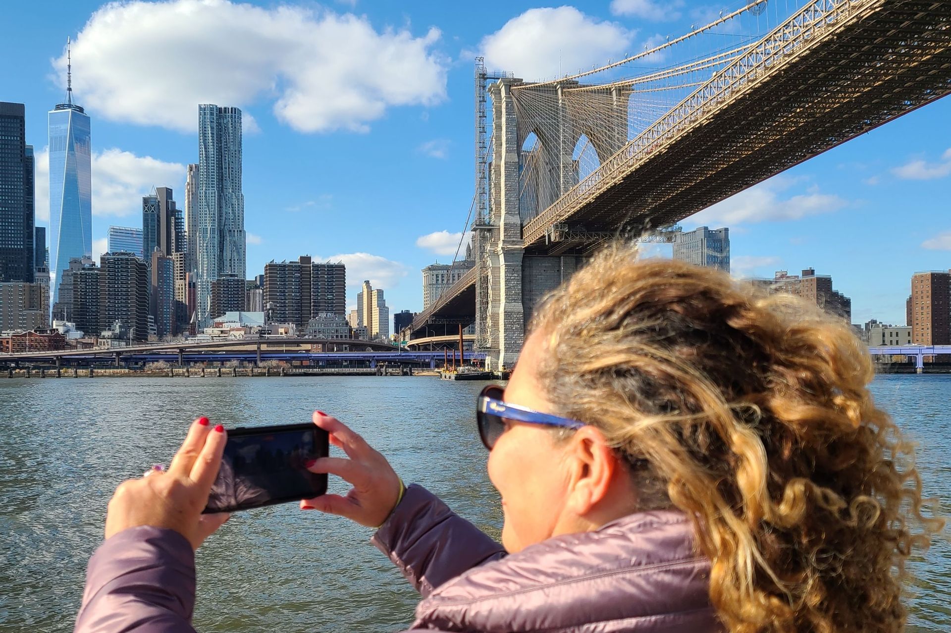 Woman taking photo of NYC skyline and Brooklyn Bridge on a sunny day.