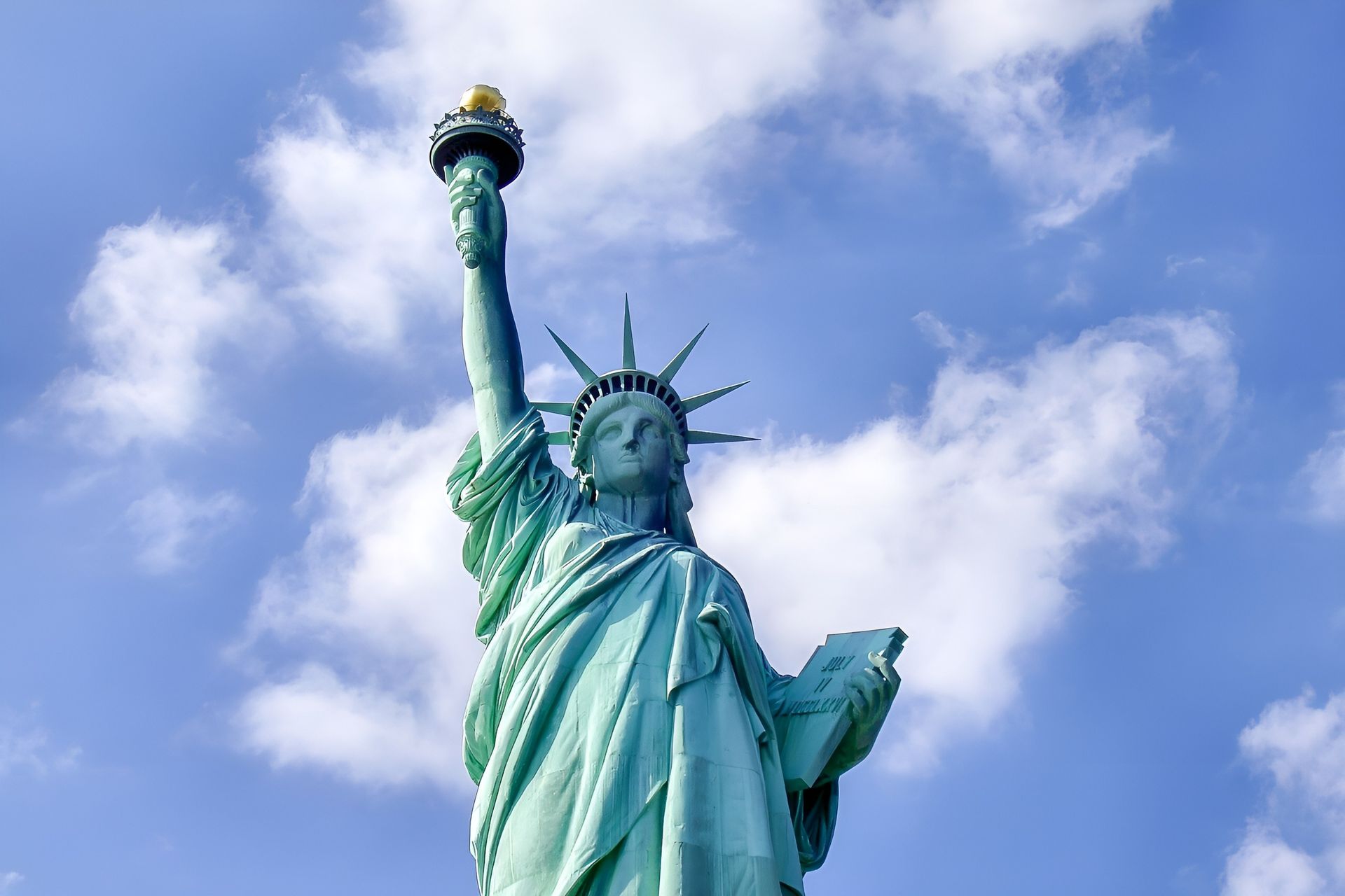 Statue of Liberty, green statue holding a torch against a blue sky with fluffy clouds.