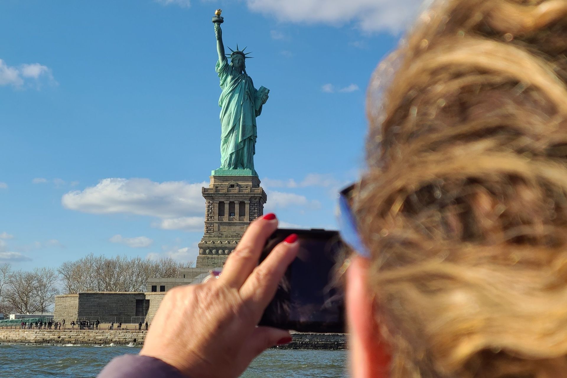 Woman taking photo of Statue of Liberty with a smartphone against a blue sky.