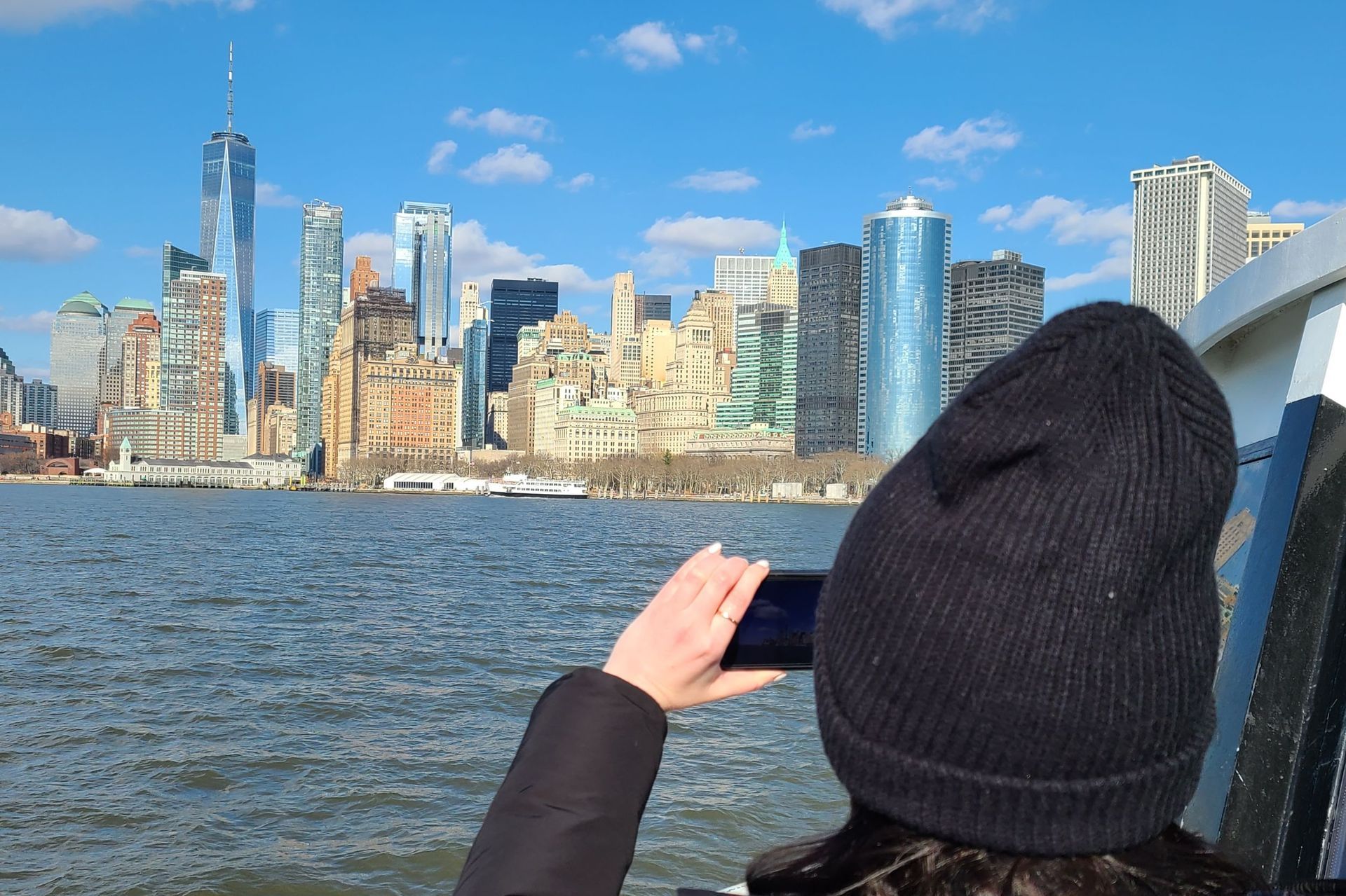 Person in black hat taking photo of NYC skyline over water on a sunny day.