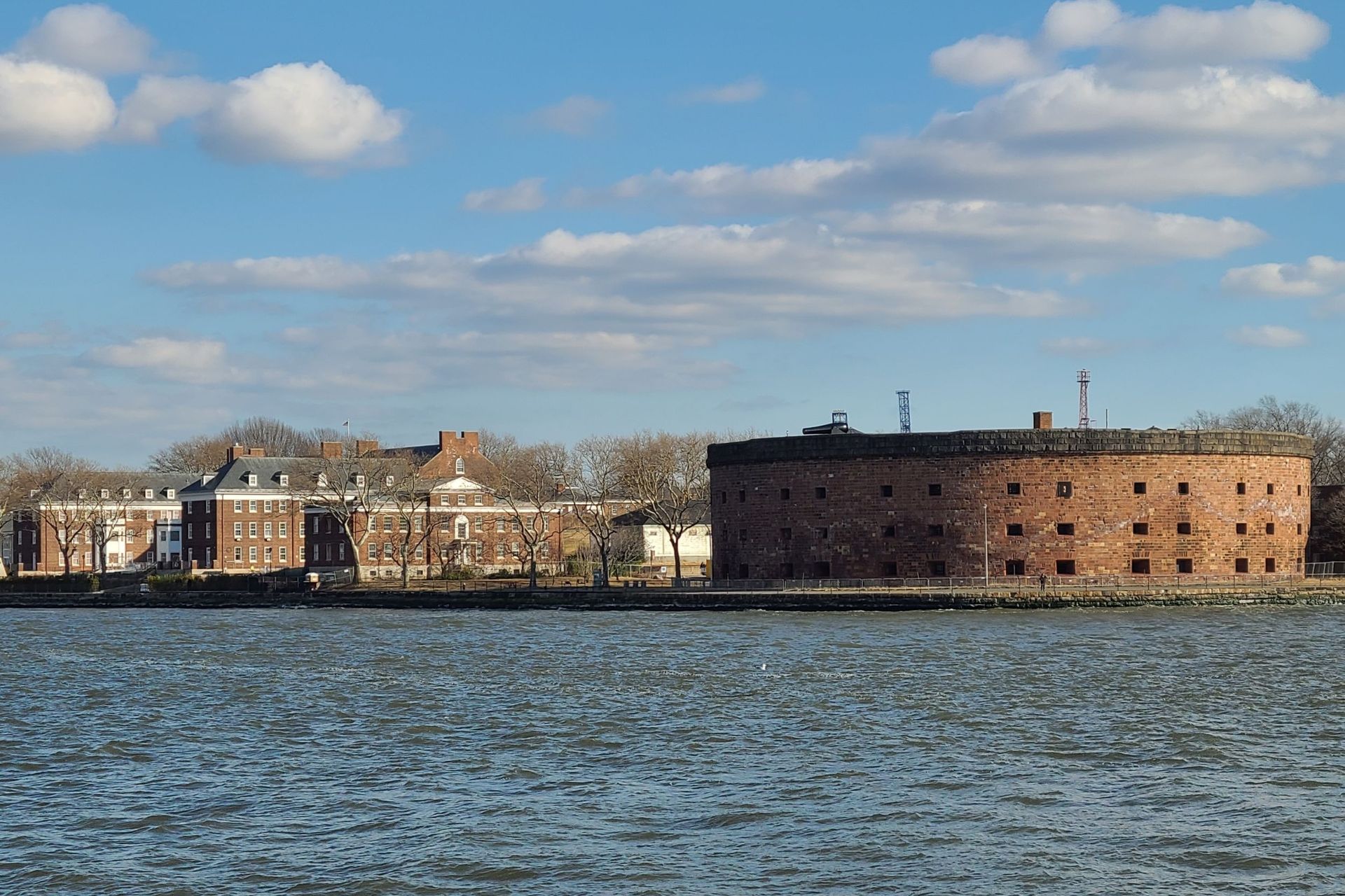 Water view of a brick, circular fort and a long, light-colored building under a blue sky.