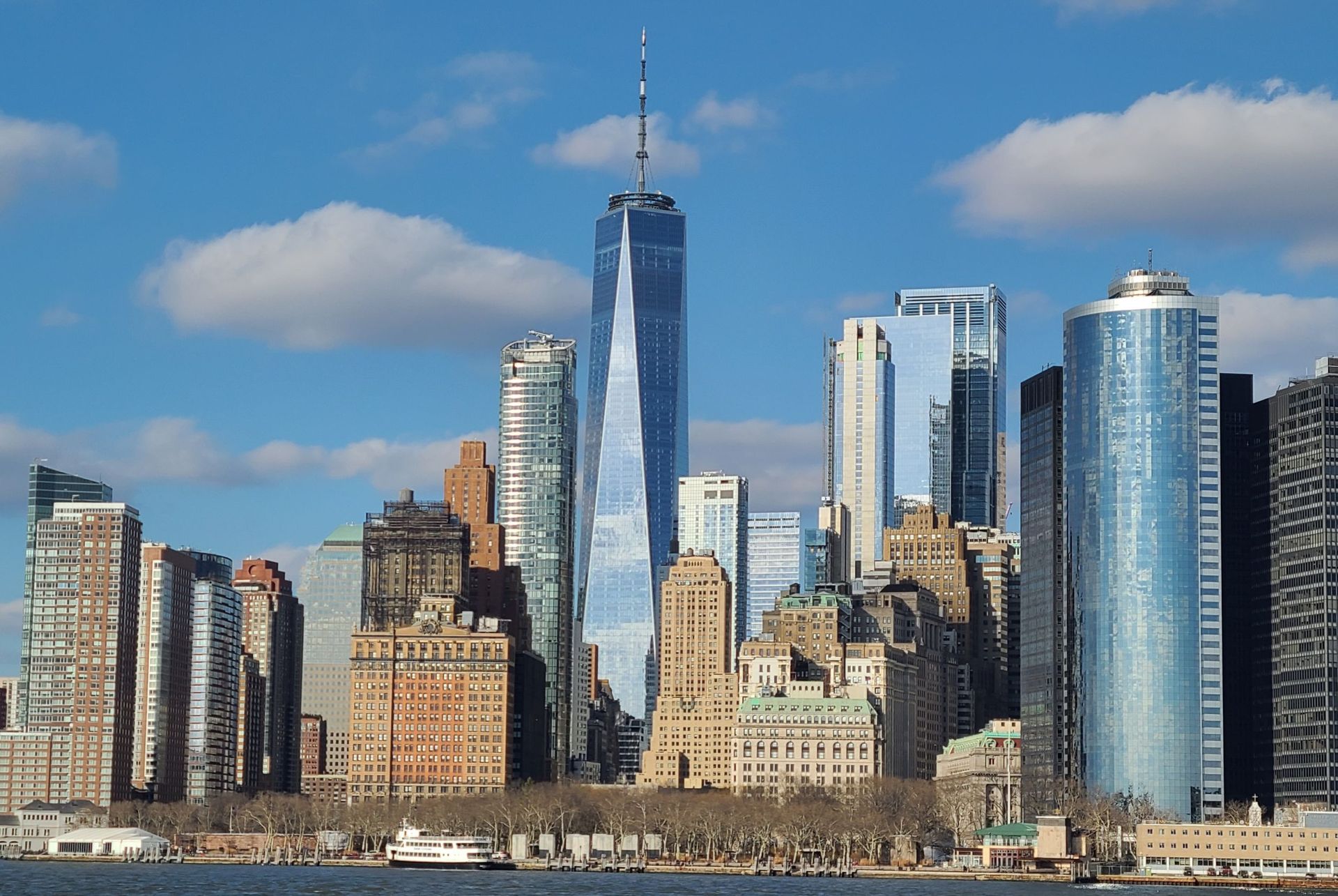 Skyline of New York City with One World Trade Center in the center, blue sky, and water in the foreground.