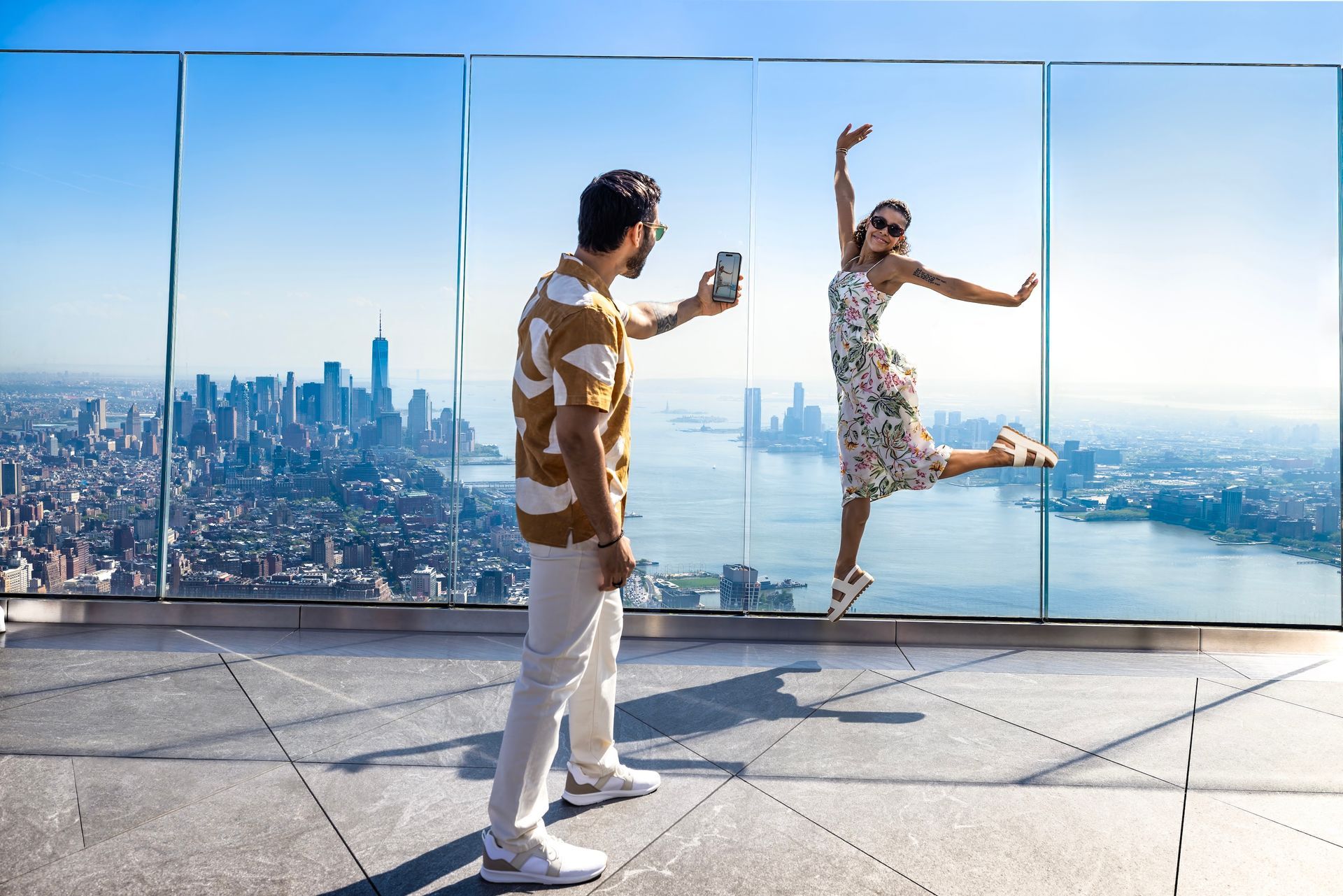 Man taking photo of woman jumping in front of city skyline, both smiling, outdoor.