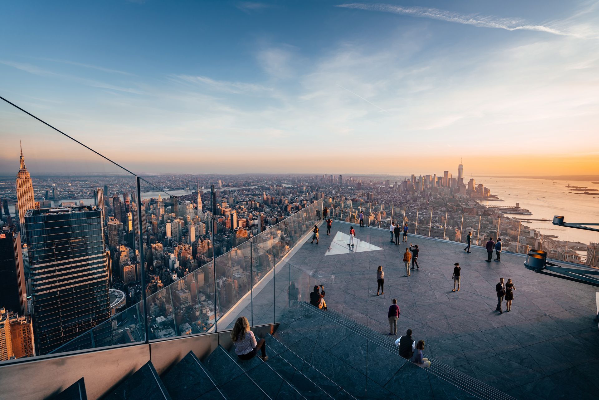 City skyline at sunset, people on observation deck, glass barrier, skyscrapers, golden light.