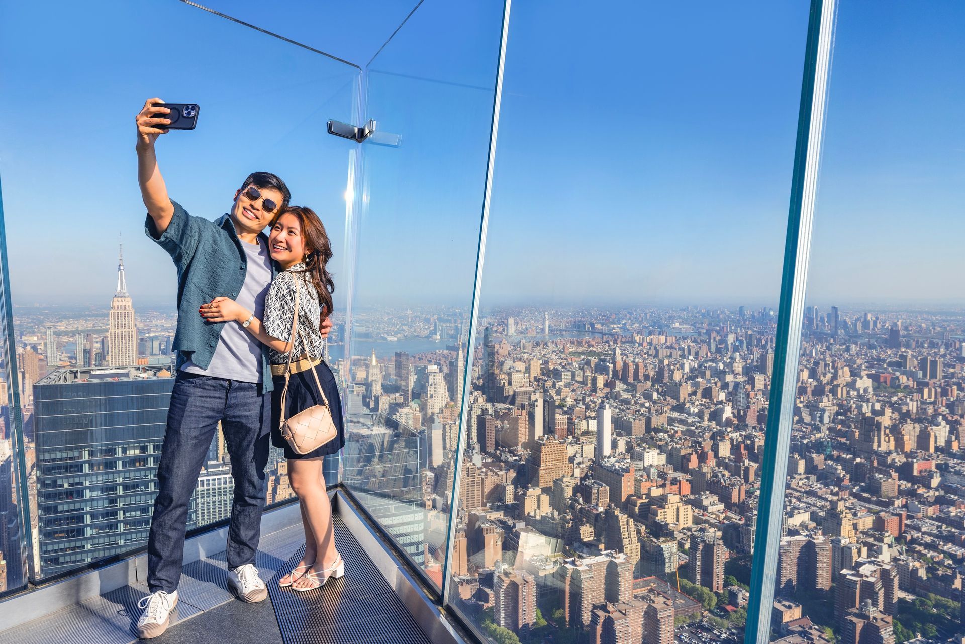Couple takes selfie on a glass platform high above a sprawling city. Blue sky and city landscape are in background.