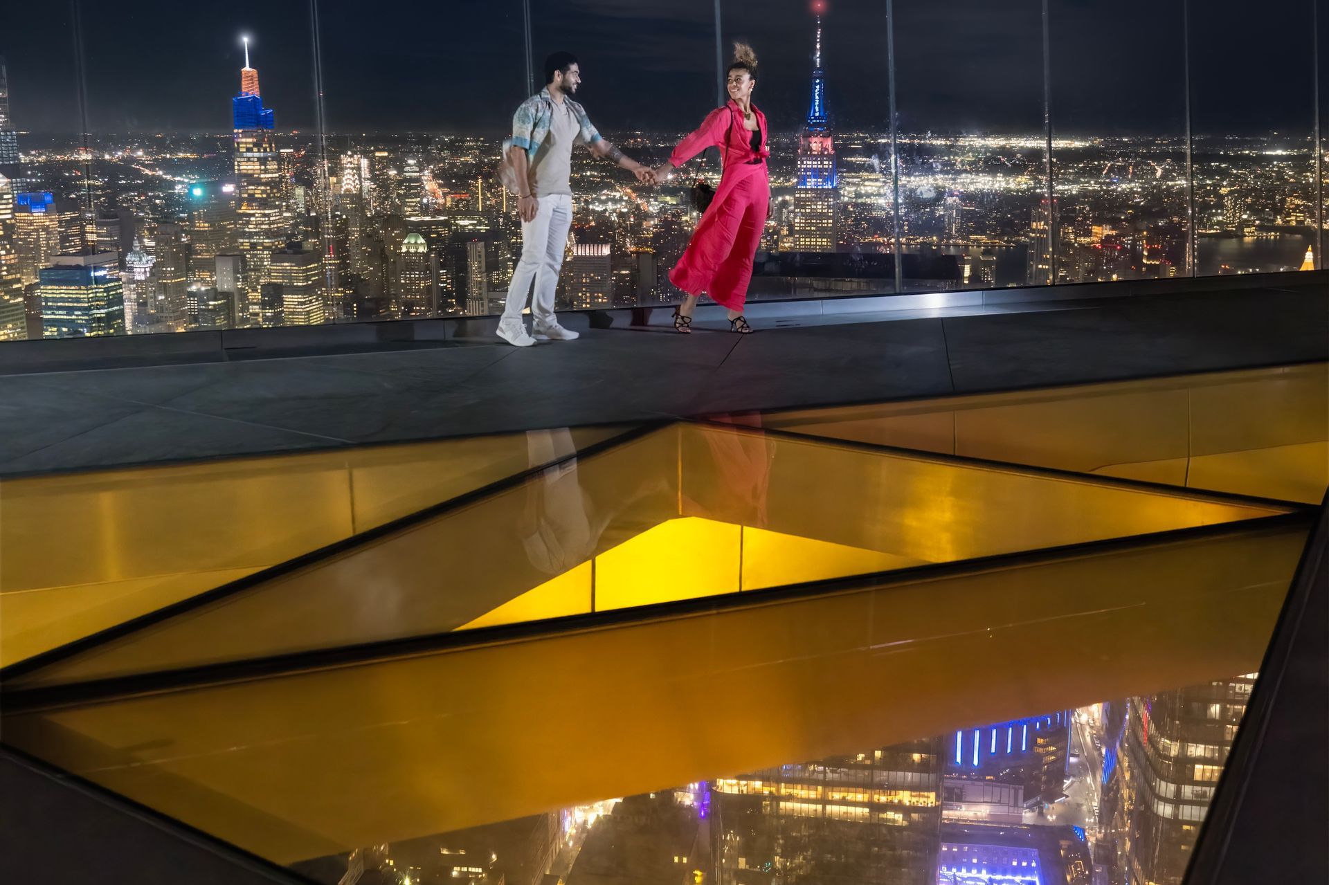 Couple holding hands on a rooftop overlooking a brightly lit city at night; a glass floor reflects the scene.