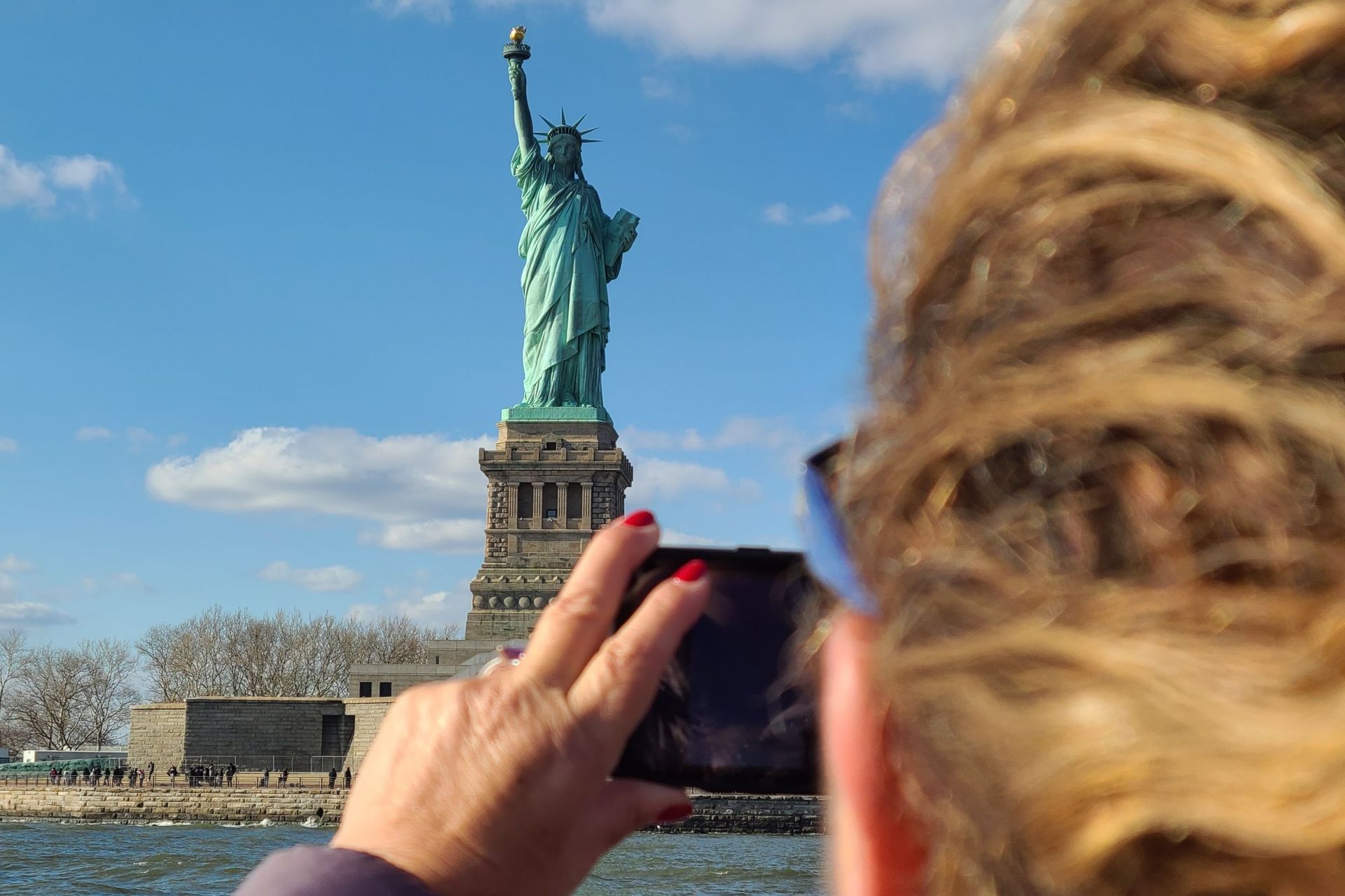 Person taking a photo of the Statue of Liberty on a sunny day.