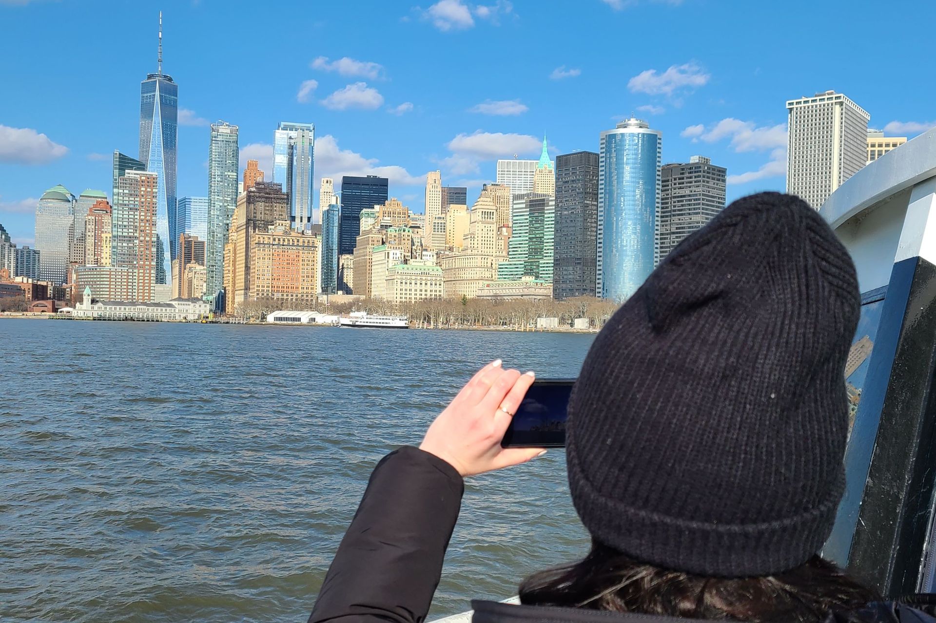 Person in black hat takes a photo of New York City skyline from a boat on a sunny day.