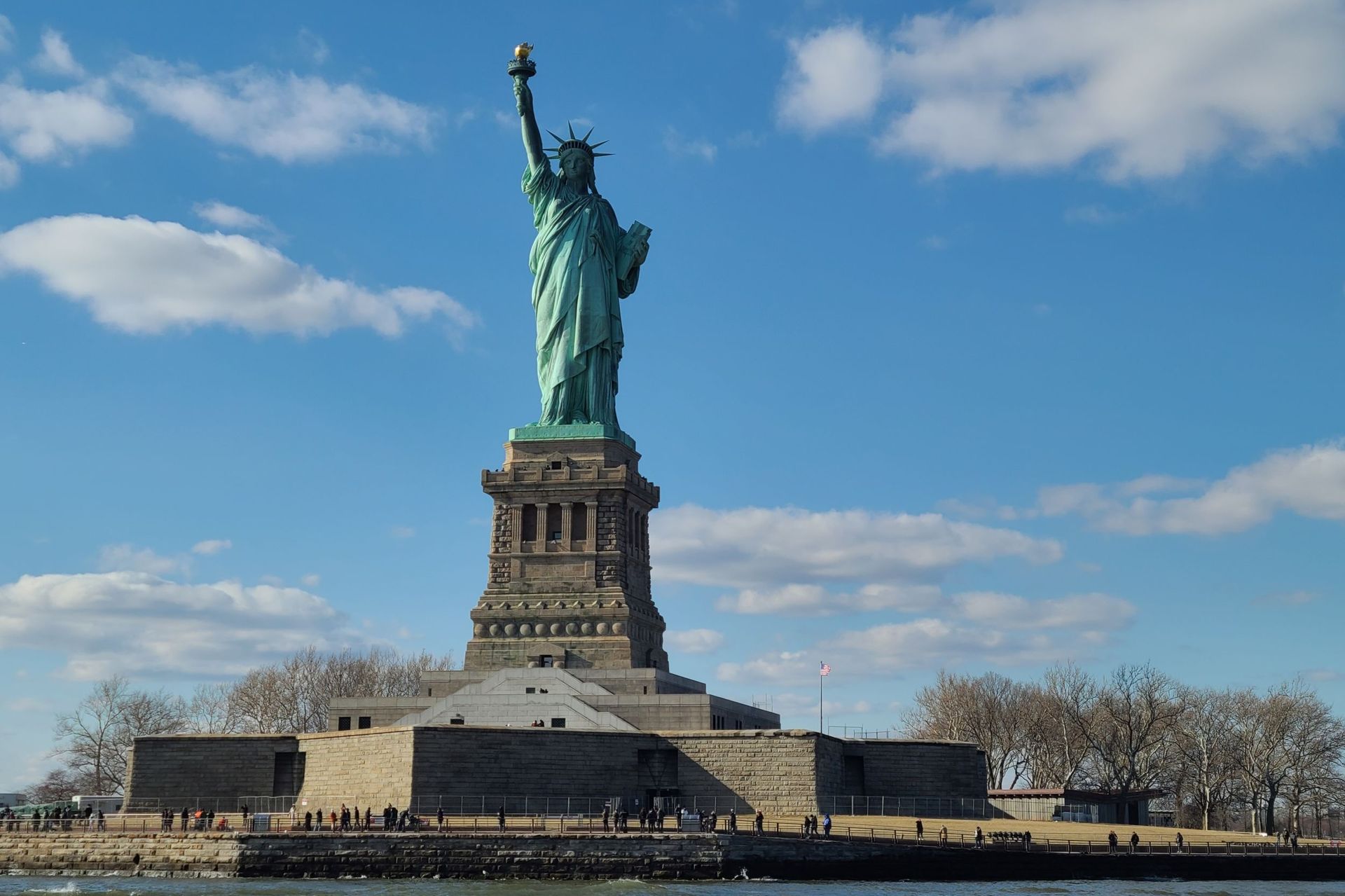 Statue of Liberty stands tall on Liberty Island against a blue sky with clouds, water in the foreground.