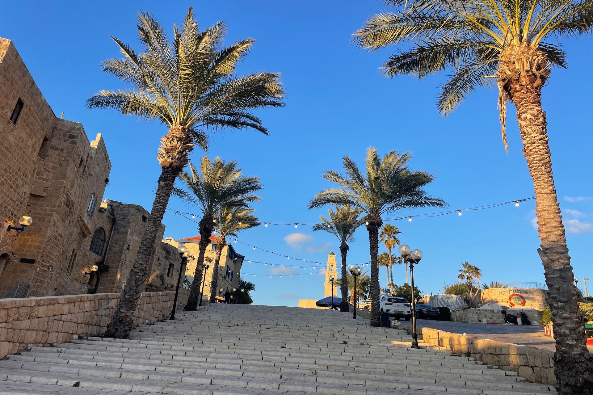 Stone steps lead uphill toward a clock tower in Jaffa, Israel, lined with palm trees under a blue sky.