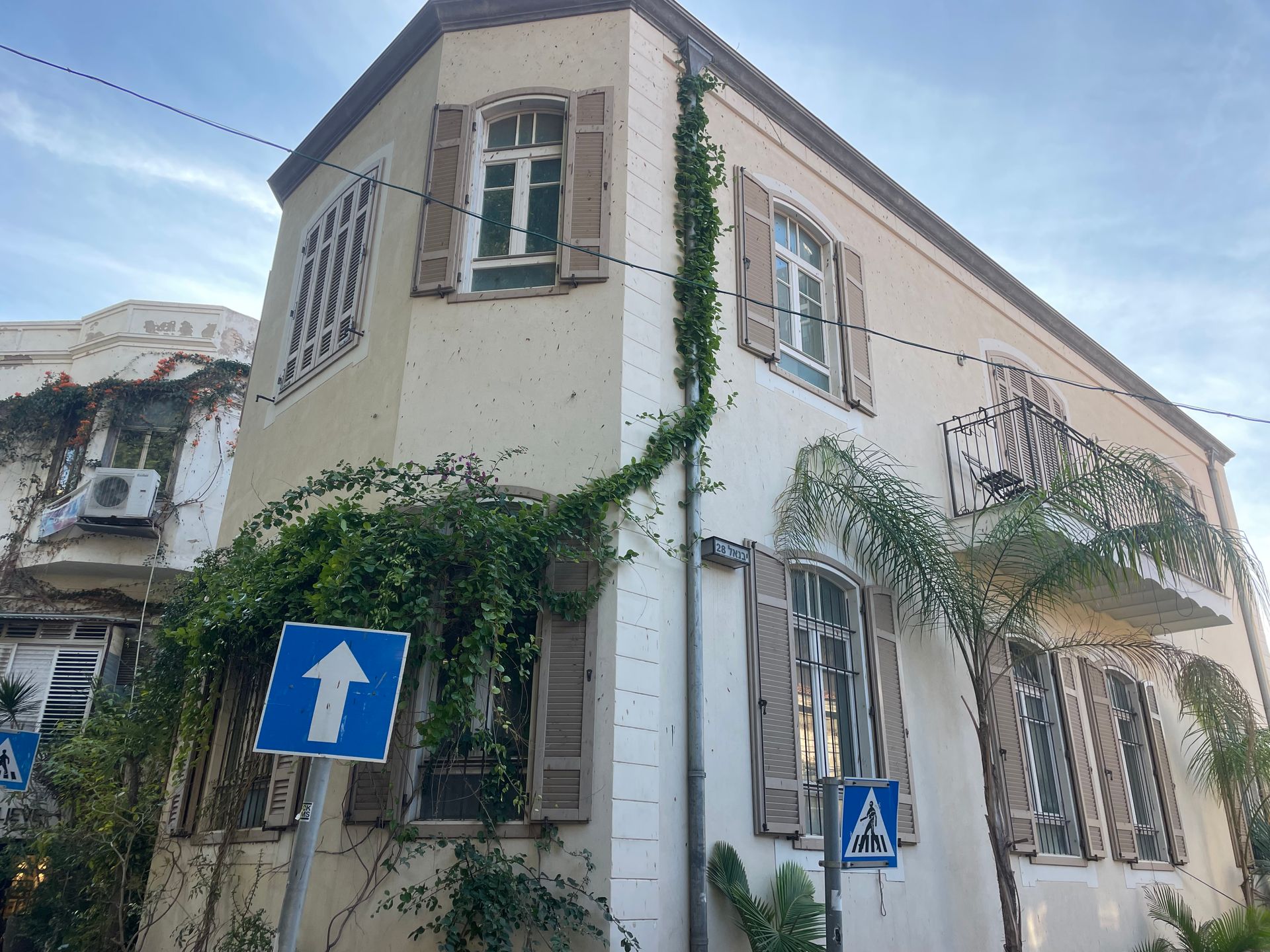 Two-story white corner building with open shutters, surrounded by climbing plants, blue street signs in Jaffa, Israel.