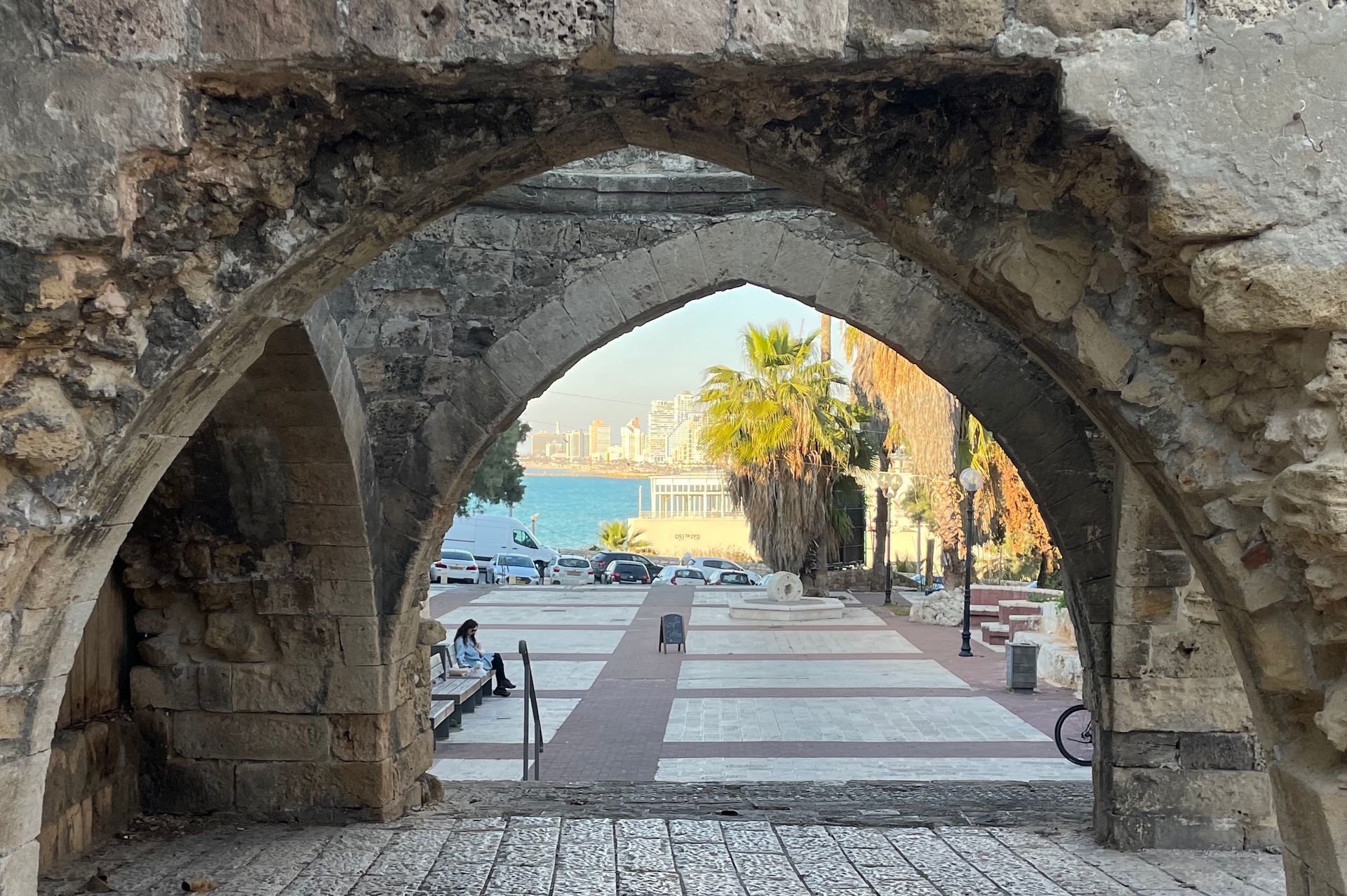 Stone archway frames a view of a seaside promenade with palm trees, cars, and people.