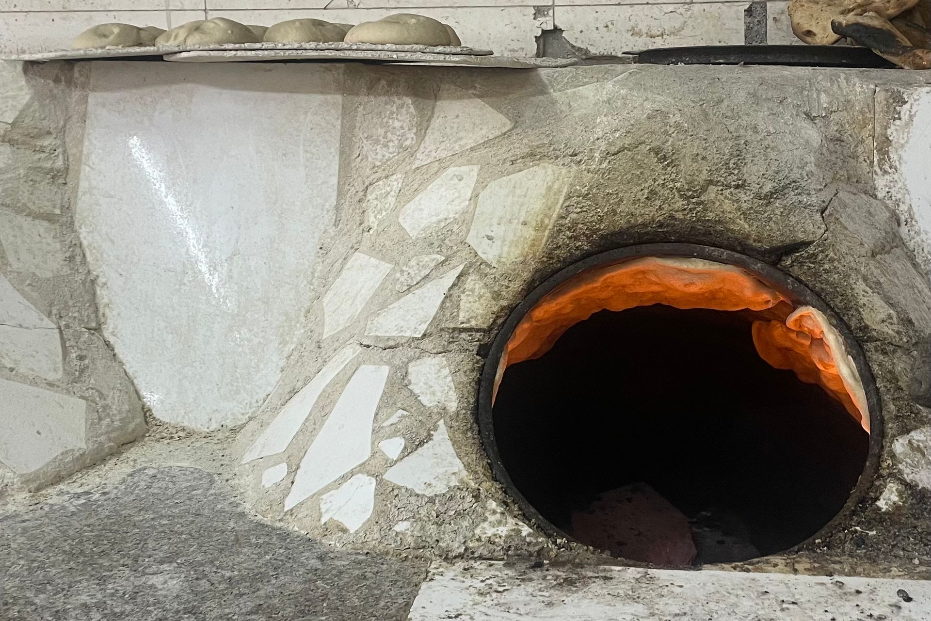 A brick oven with a round opening and orange-glowing interior. Bread on the ledge above.