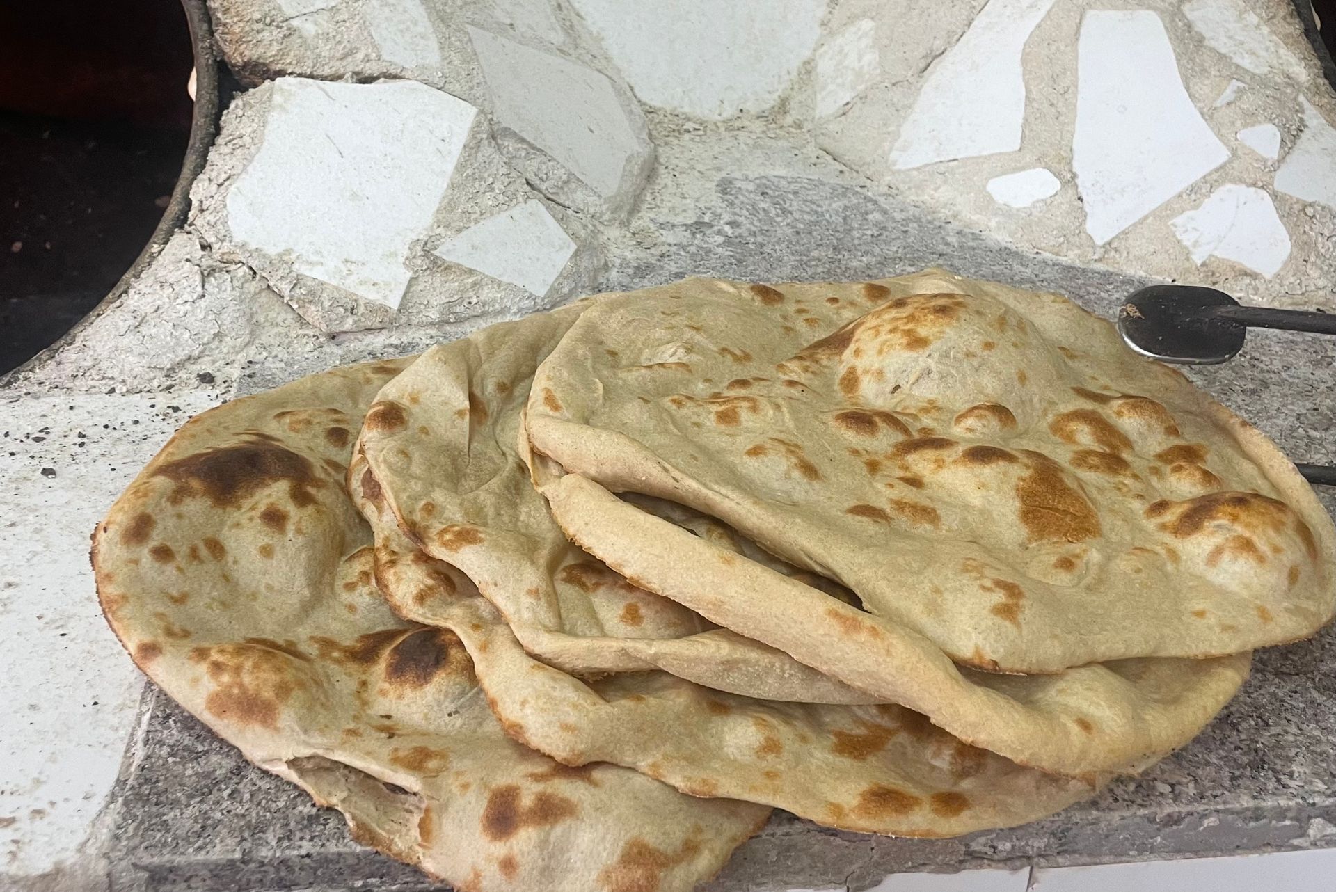 Pile of round, golden-brown flatbreads on a stone surface, near a textured oven.