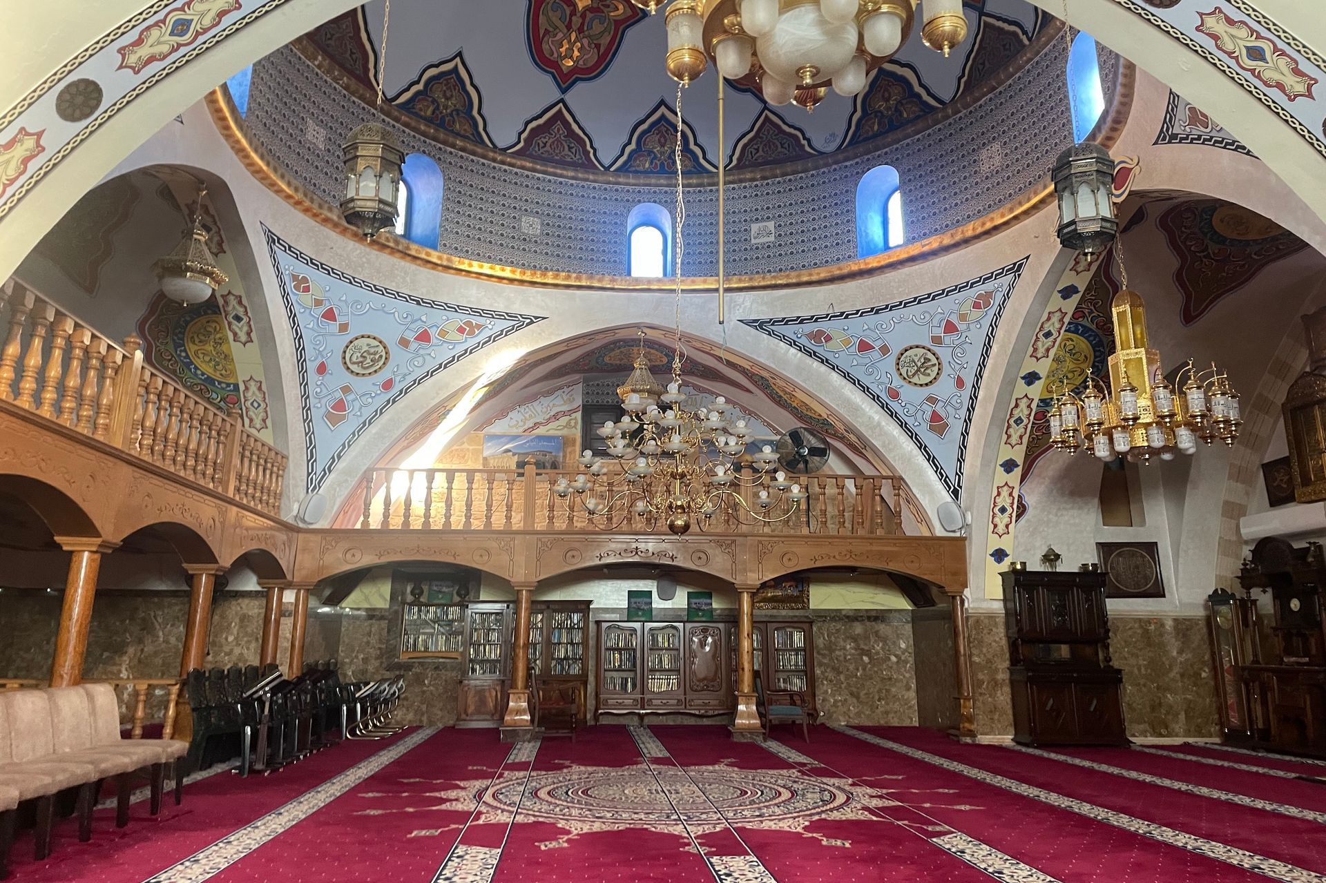 Interior view of a mosque with a large domed ceiling, patterned walls, red carpet, and chandeliers.