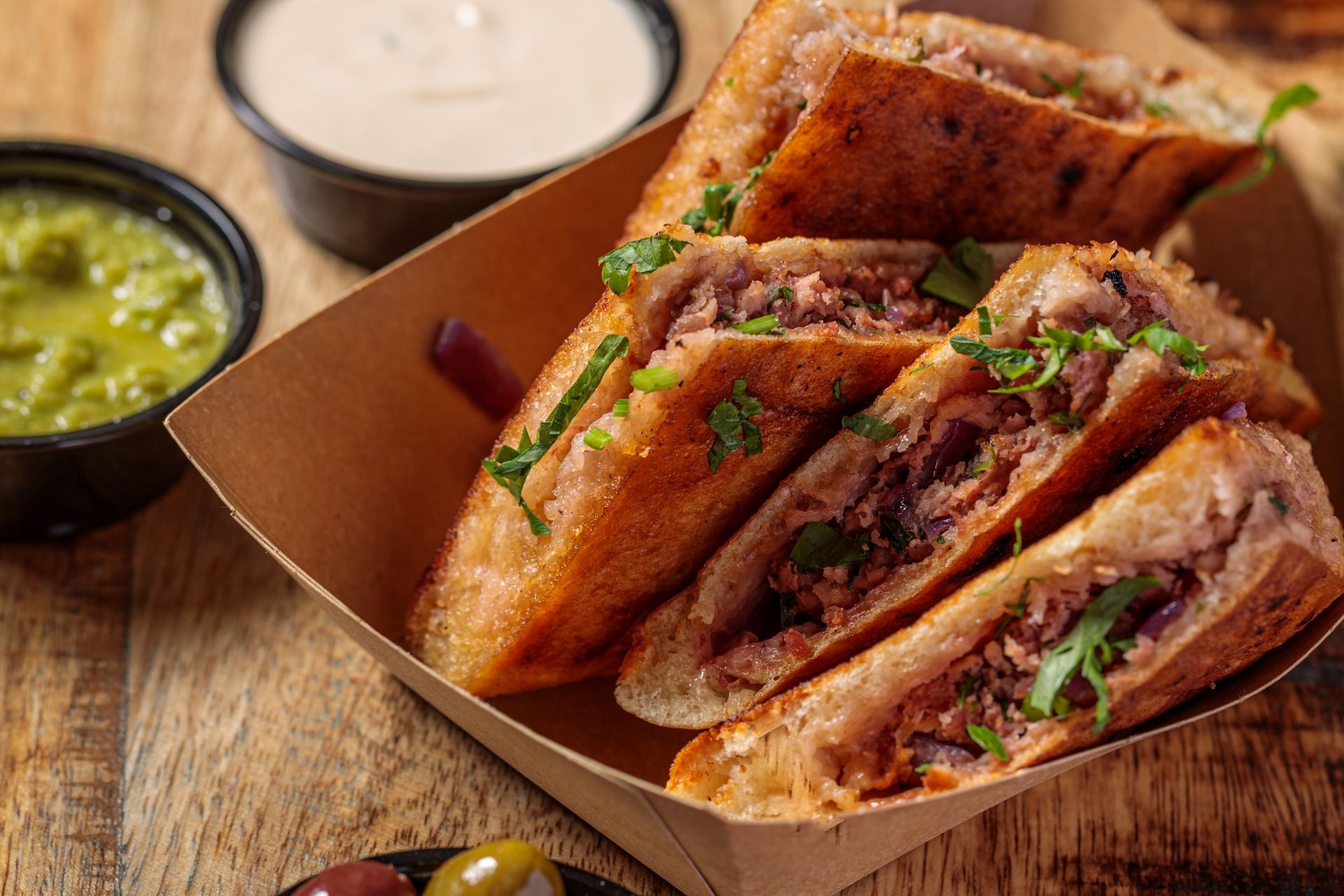 Sandwiches cut in half with meat filling, served in a paper tray with sauces and olives on a wooden surface.