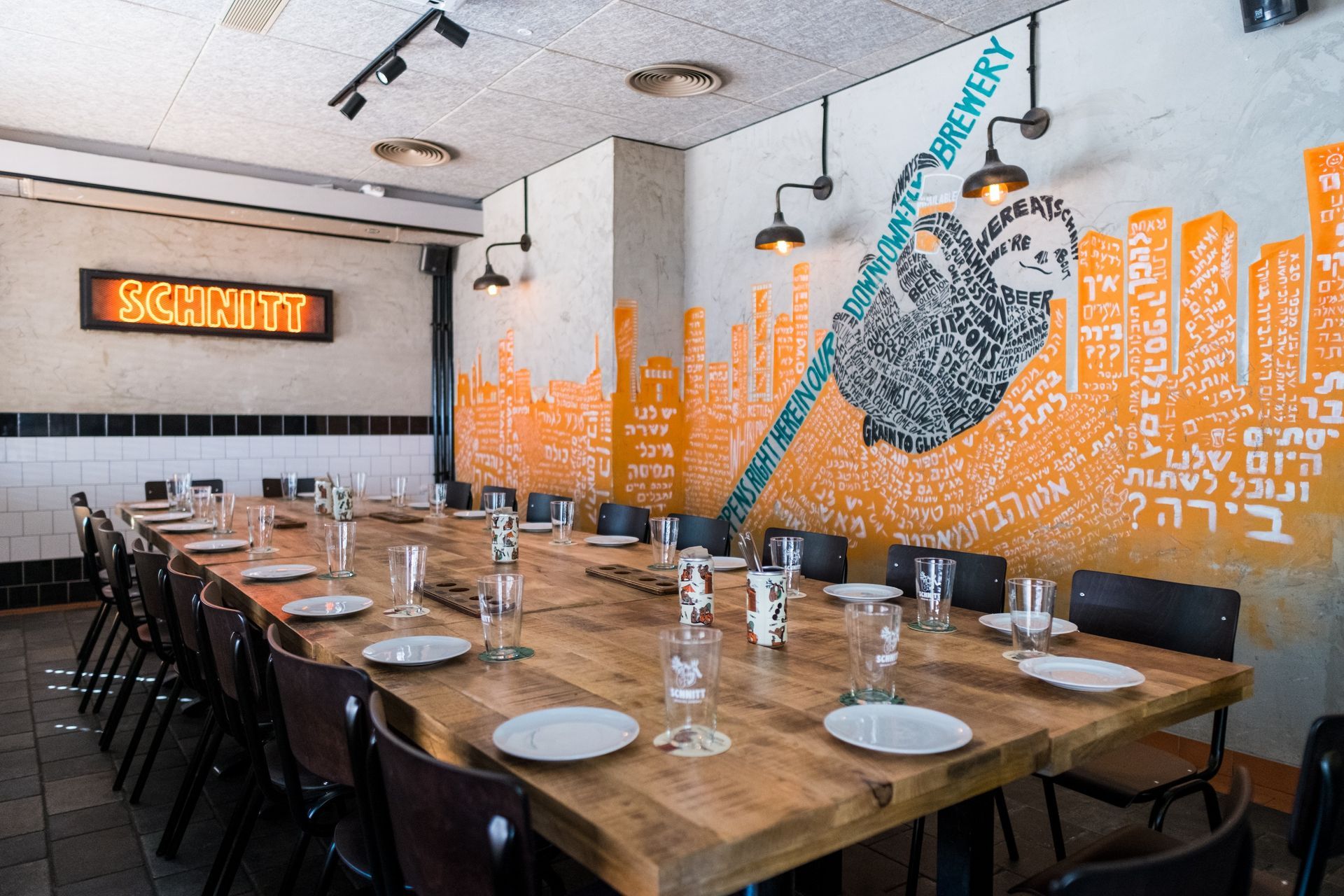 Long wooden table set for a meal, with chairs, plates, and glasses in a restaurant; wall art.