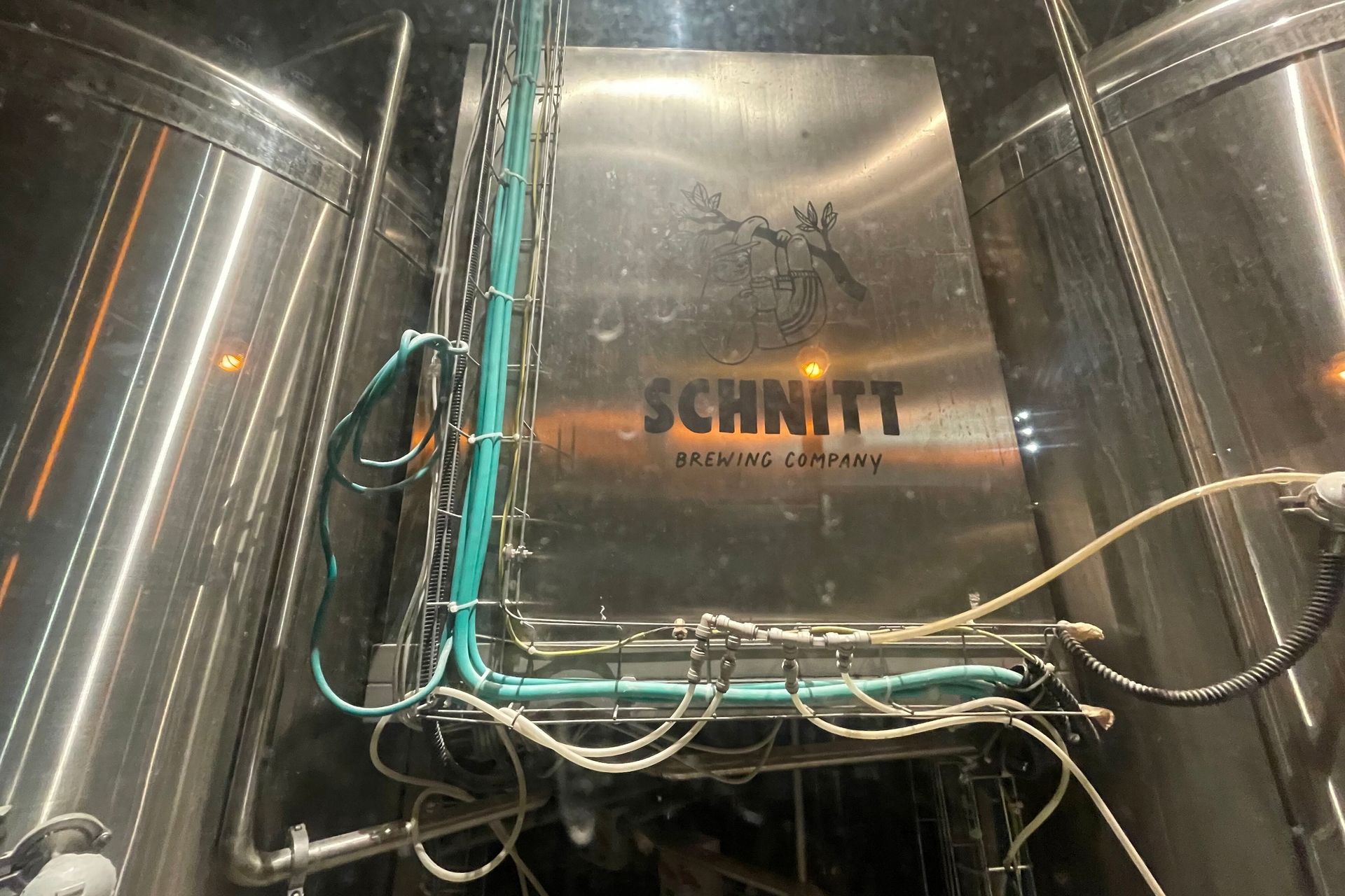 Stainless steel tanks at Schnitt Brewing Company, with central panel featuring brewery logo and green and white tubing.
