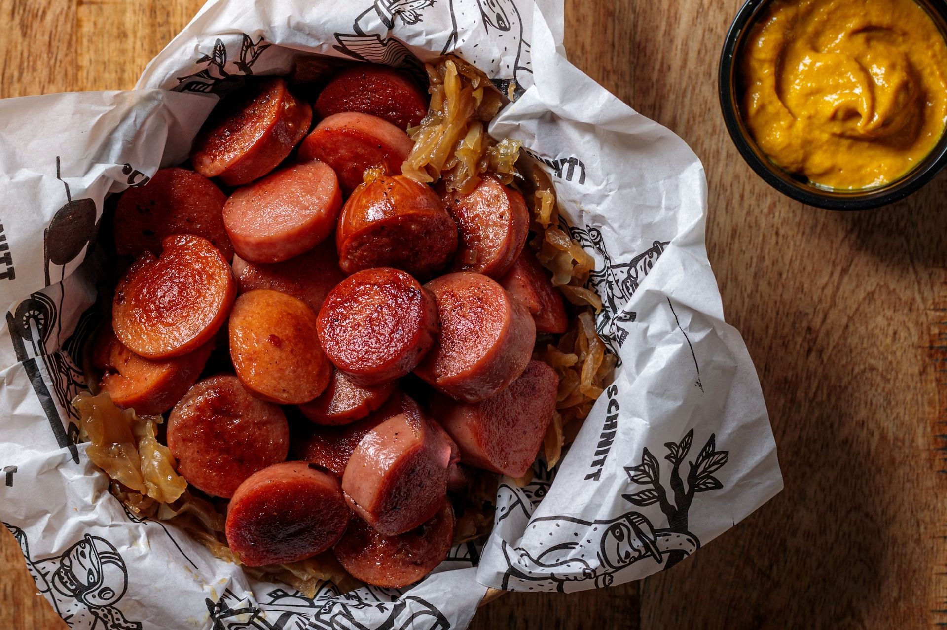 Sausage rounds with caramelized onions, served with mustard on a wooden table in a paper-lined basket.