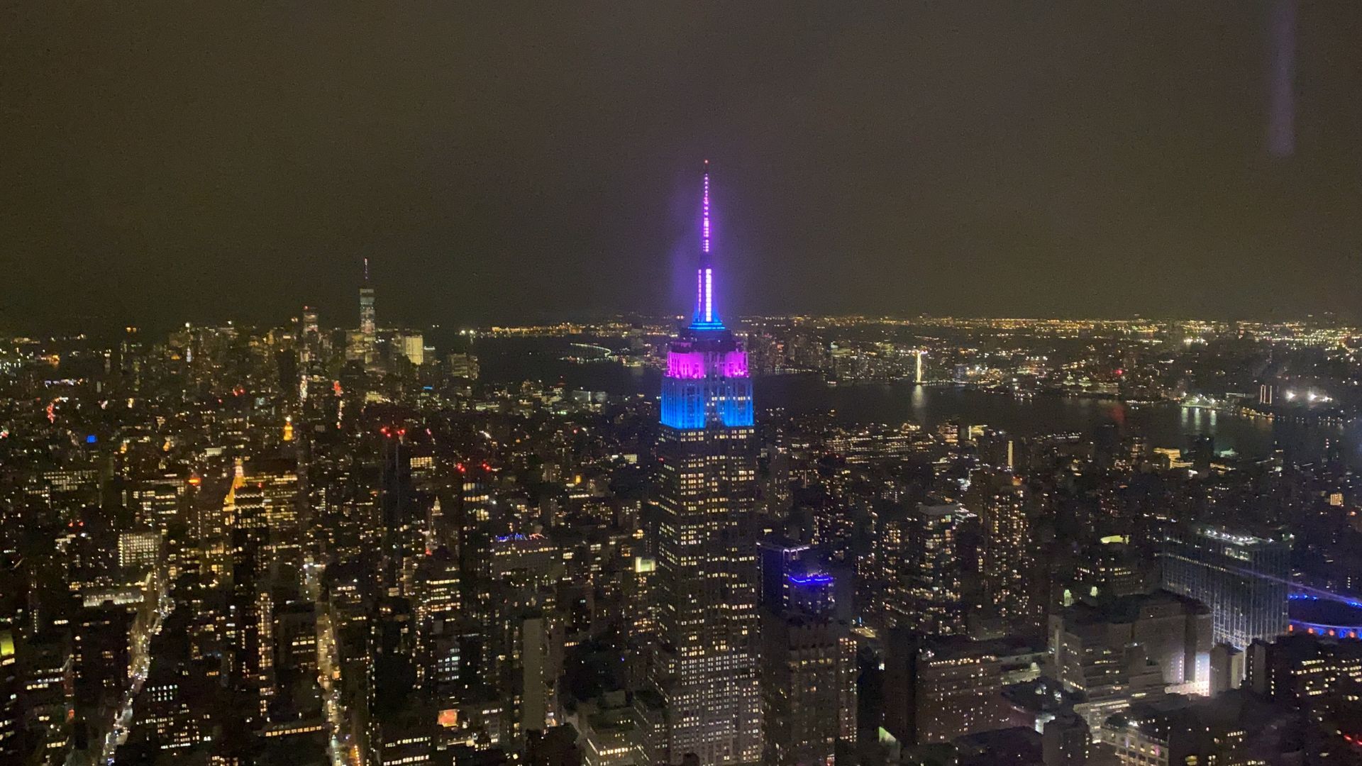 Night view of the Empire State Building lit in purple, blue, and pink over a brightly lit cityscape.