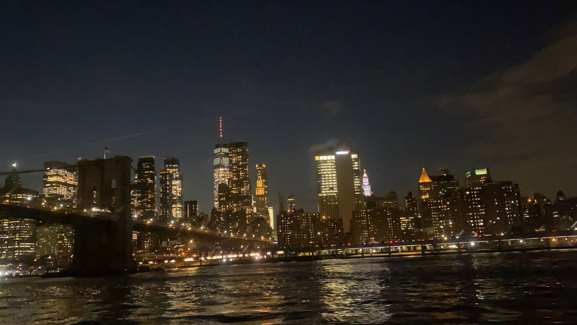 Nighttime view of the New York City skyline, with lit buildings and the Brooklyn Bridge. Lights reflect on the water.