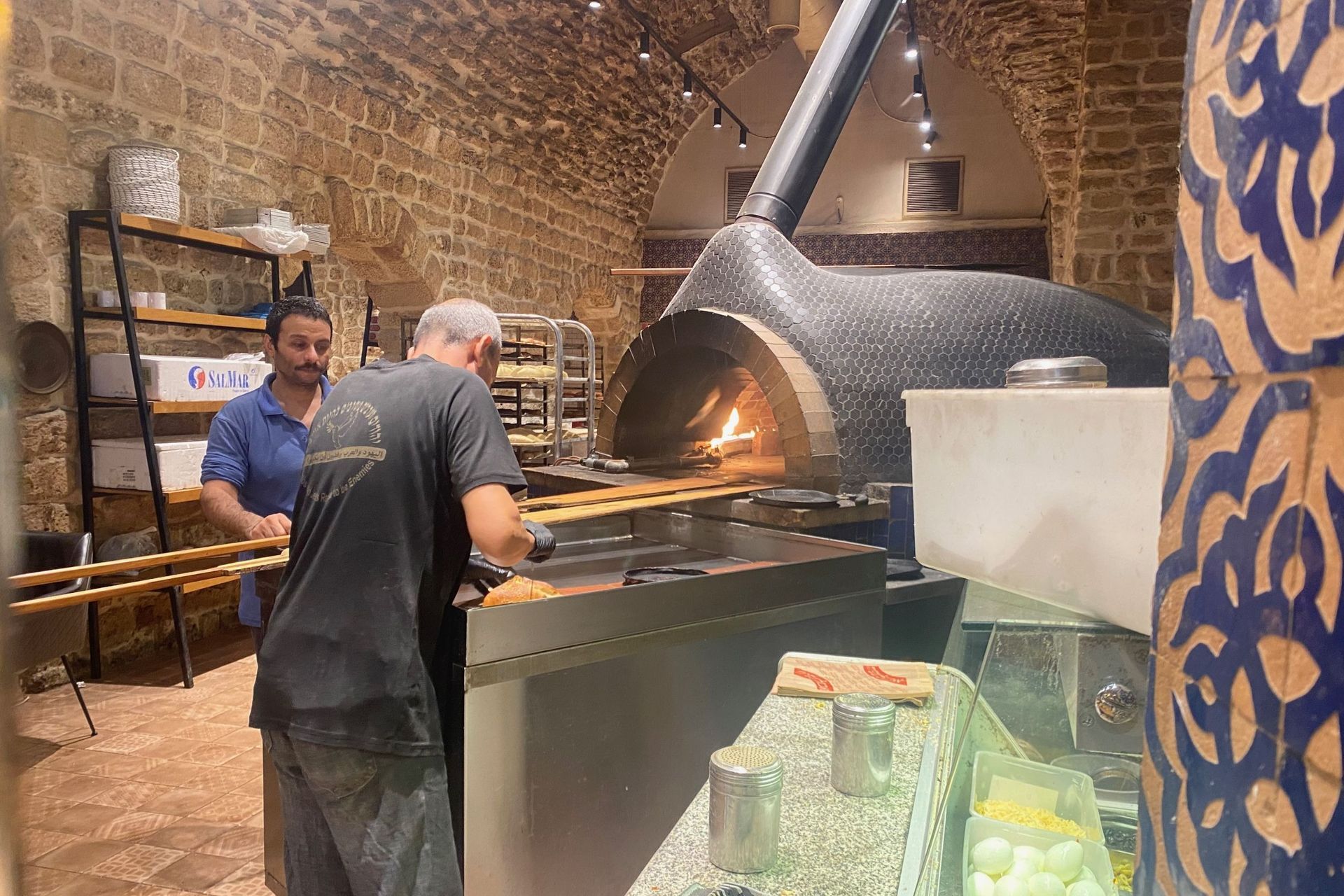 Two men preparing food near a large, lit oven in a stone-walled bakery.