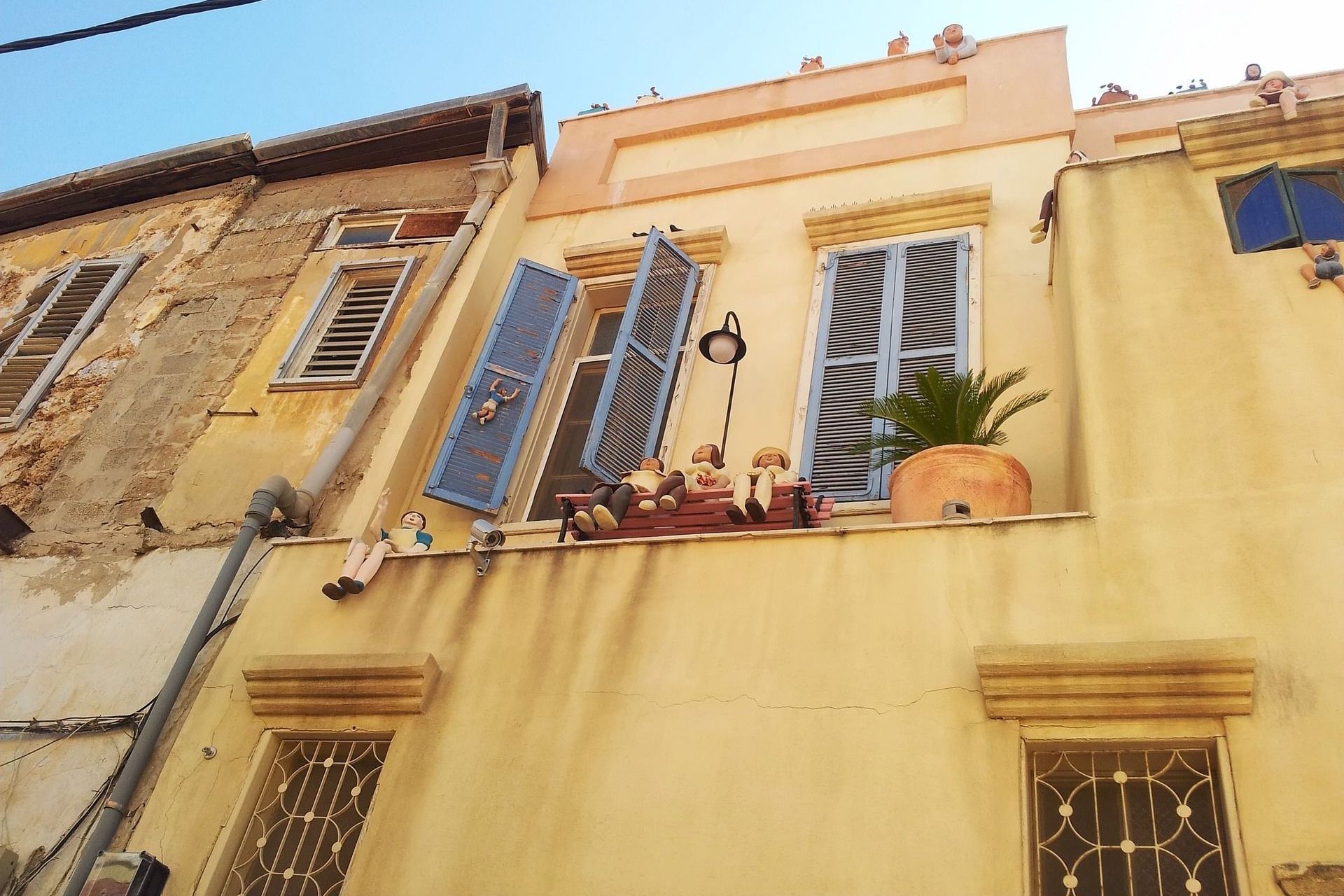 Two-story yellow building with blue shutters open, balcony with decorations, and a sunny day.