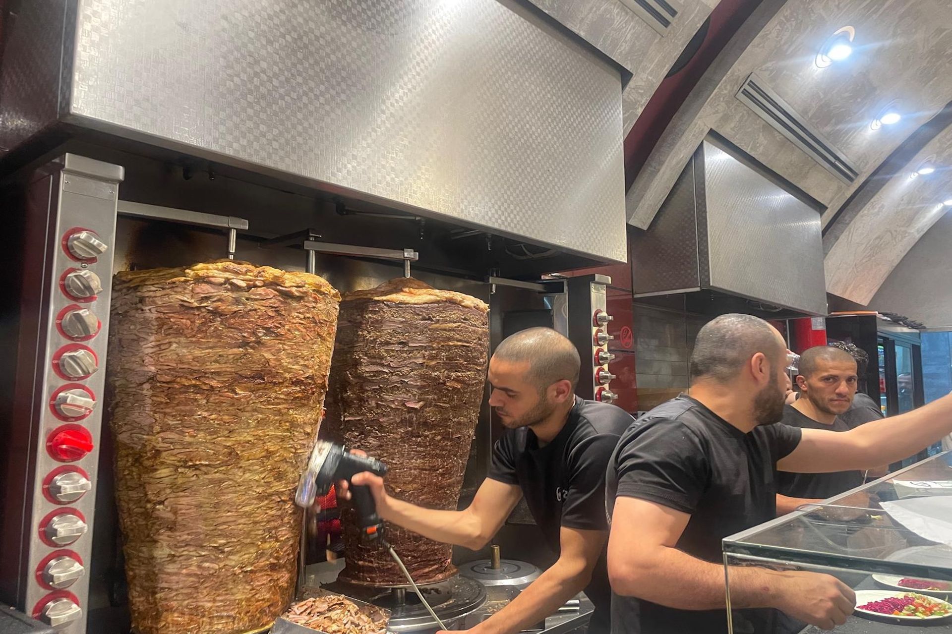 Three men preparing shawarma at a restaurant, with stacked meat roasting, and stainless steel equipment.