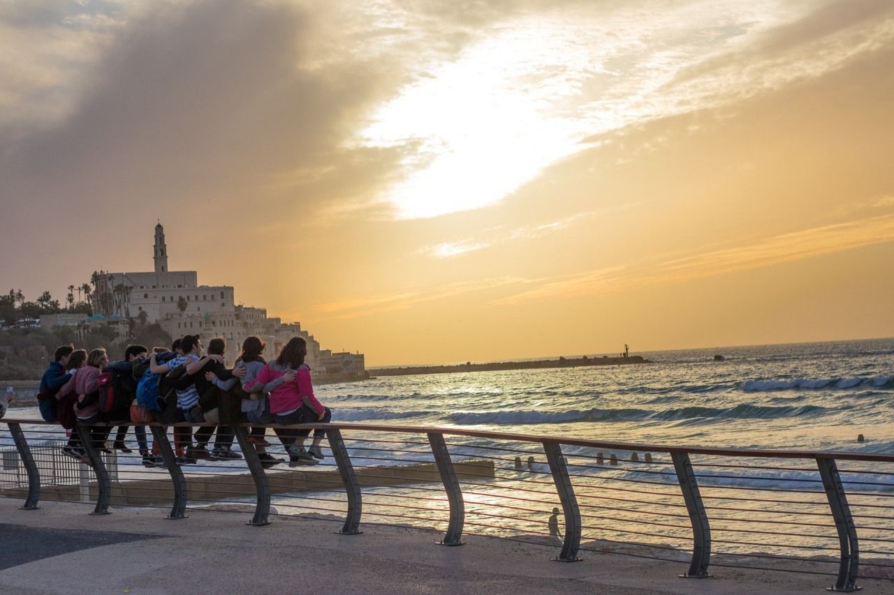 People sit on a railing, overlooking the sea at sunset near a historic building in Jaffa, Israel.