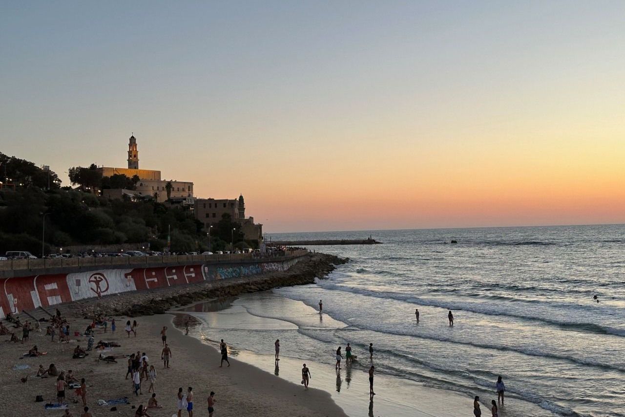 Beach at sunset with people, buildings on a hill, and colorful sky over the water.