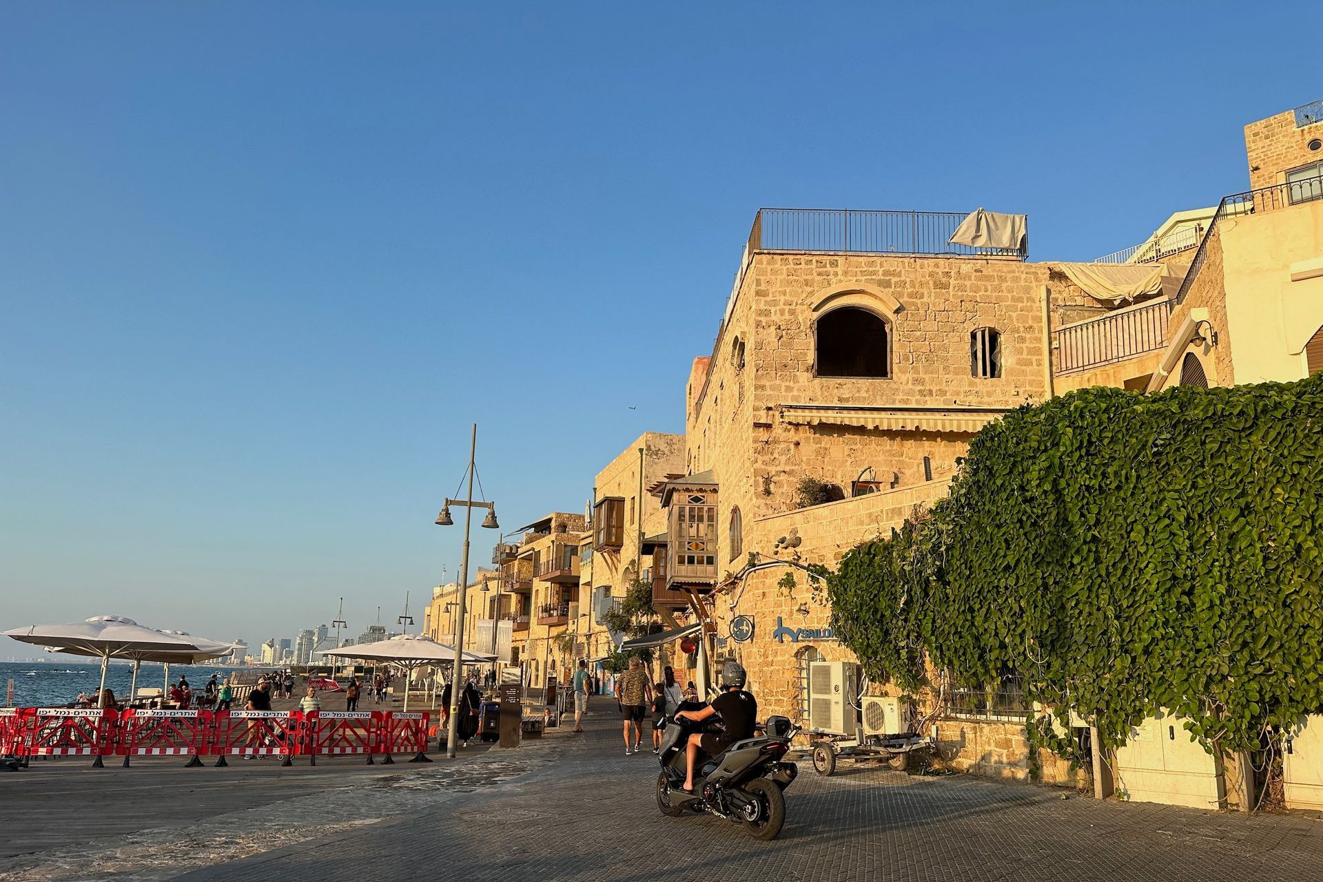 Stone buildings line a seaside promenade in Jaffa, Israel, with people, a motorcycle, and the Mediterranean Sea.
