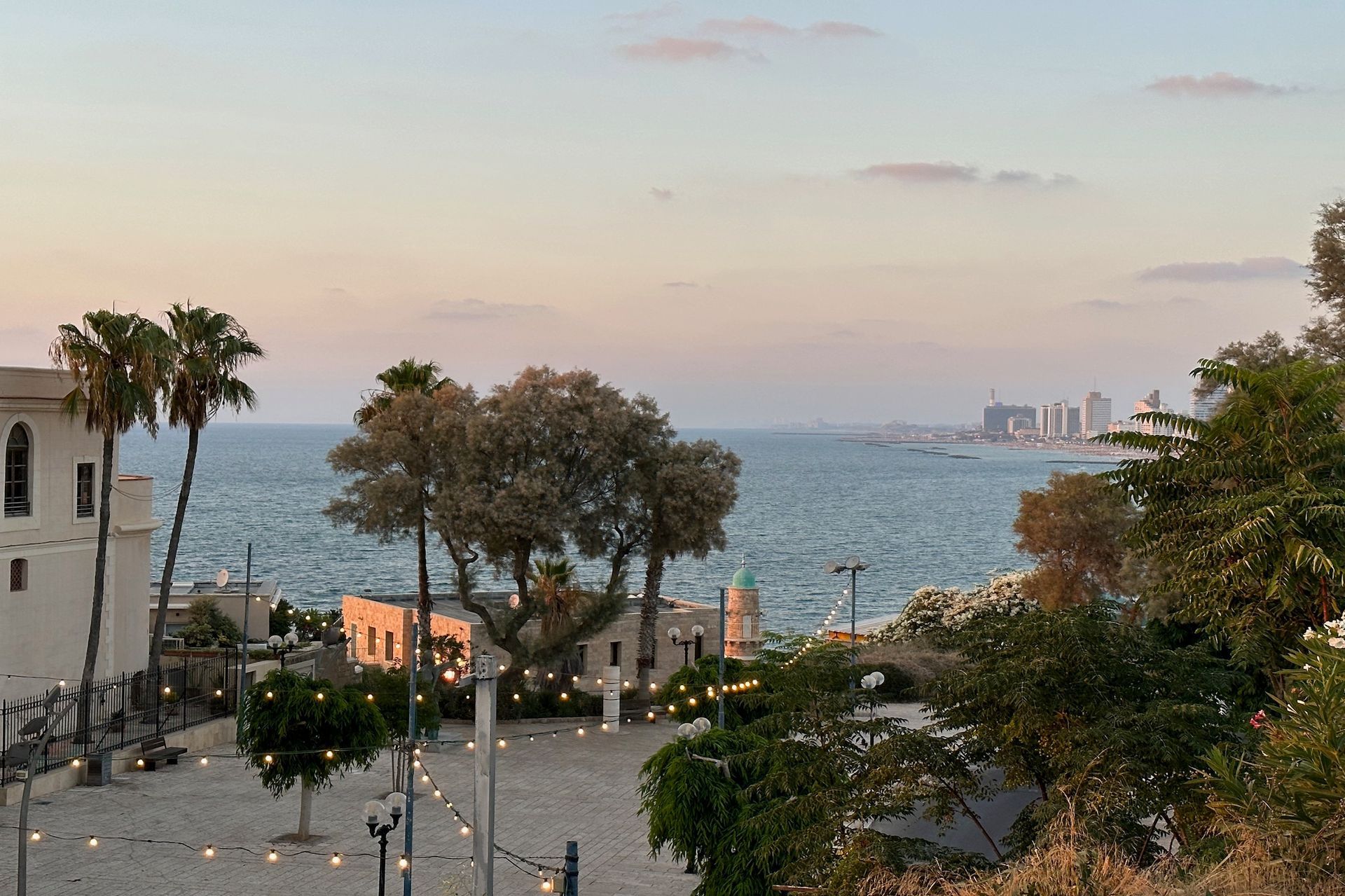 Sunset over the Mediterranean Sea from a plaza in Jaffa, Israel, with palm trees, buildings, and city skyline.