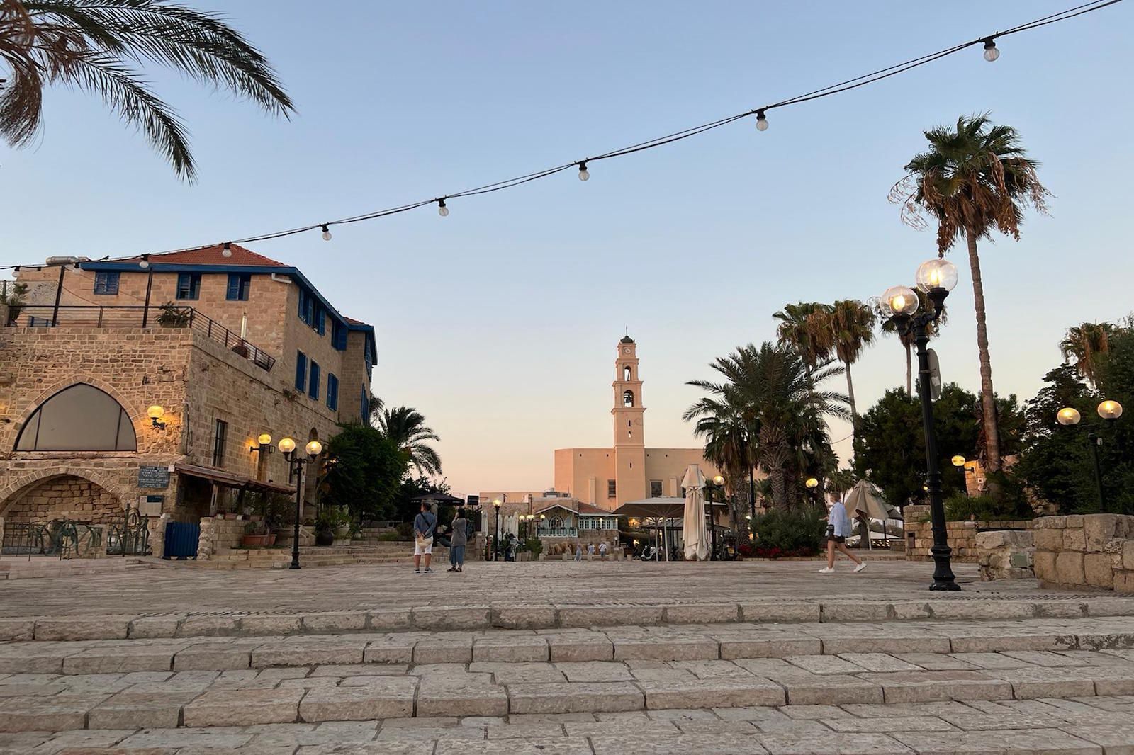 Stone steps lead to a square with a church and buildings. Dusk with palm trees and string lights.