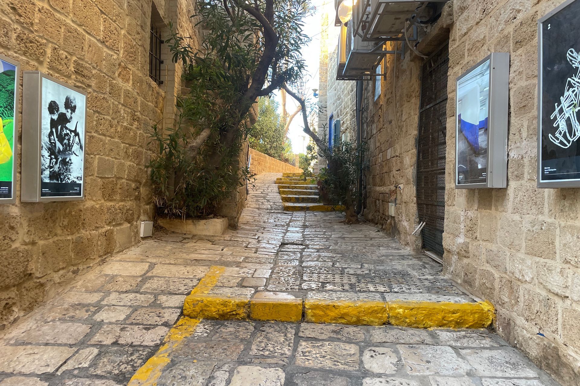Narrow cobblestone alley with stone walls and art displays. Yellow curb. Steps lead uphill.