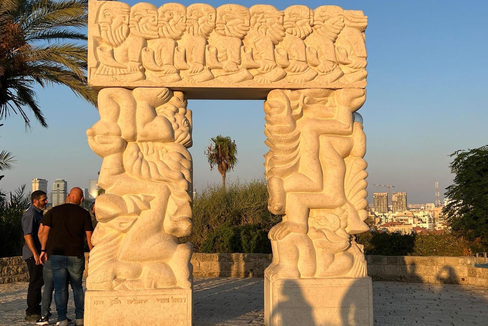 Stone archway sculpture in a park with figures, two men standing near it, city in the distance.