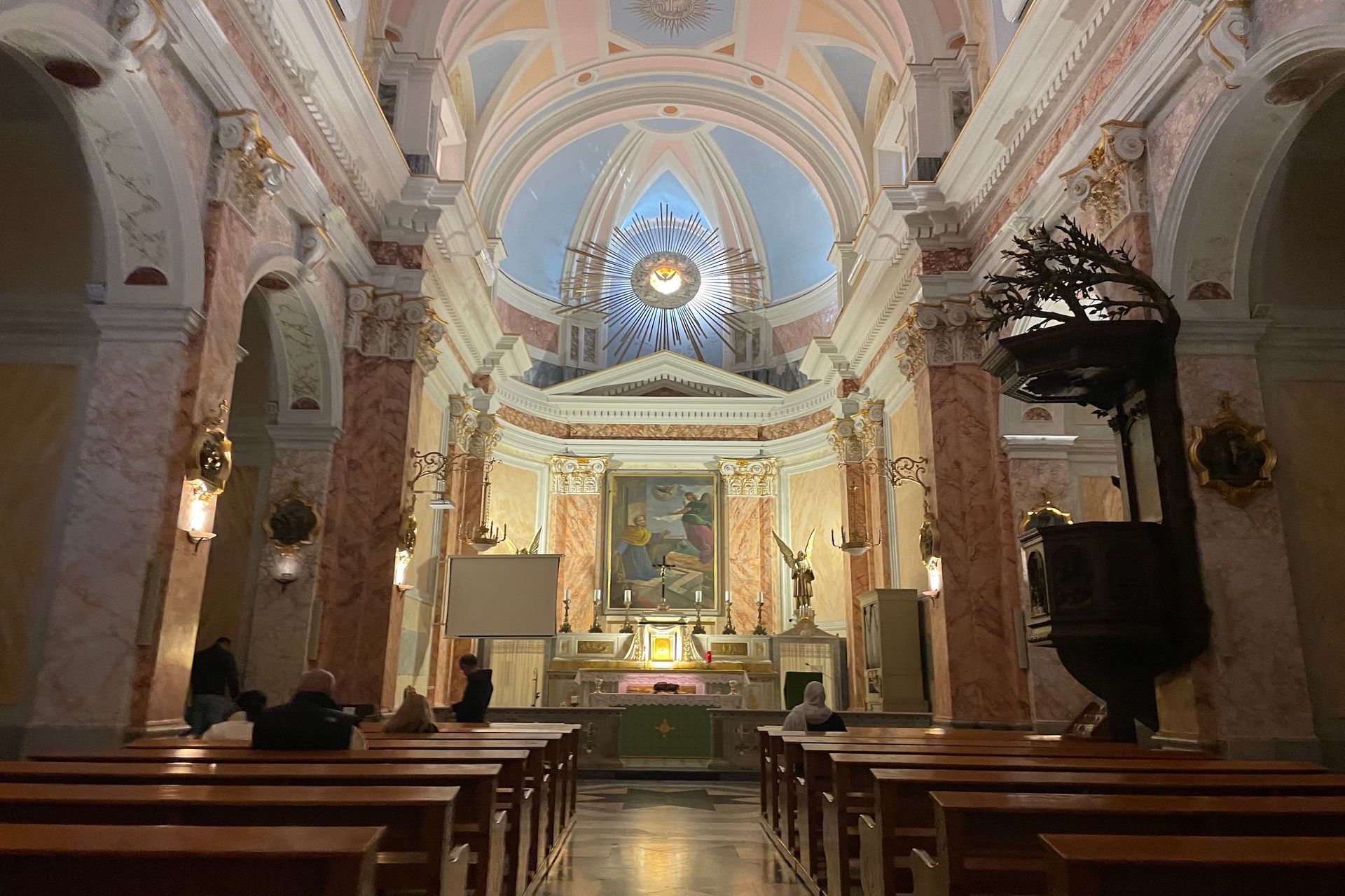 Interior view of a church with an altar, pews, and a painted ceiling, with people in the foreground.