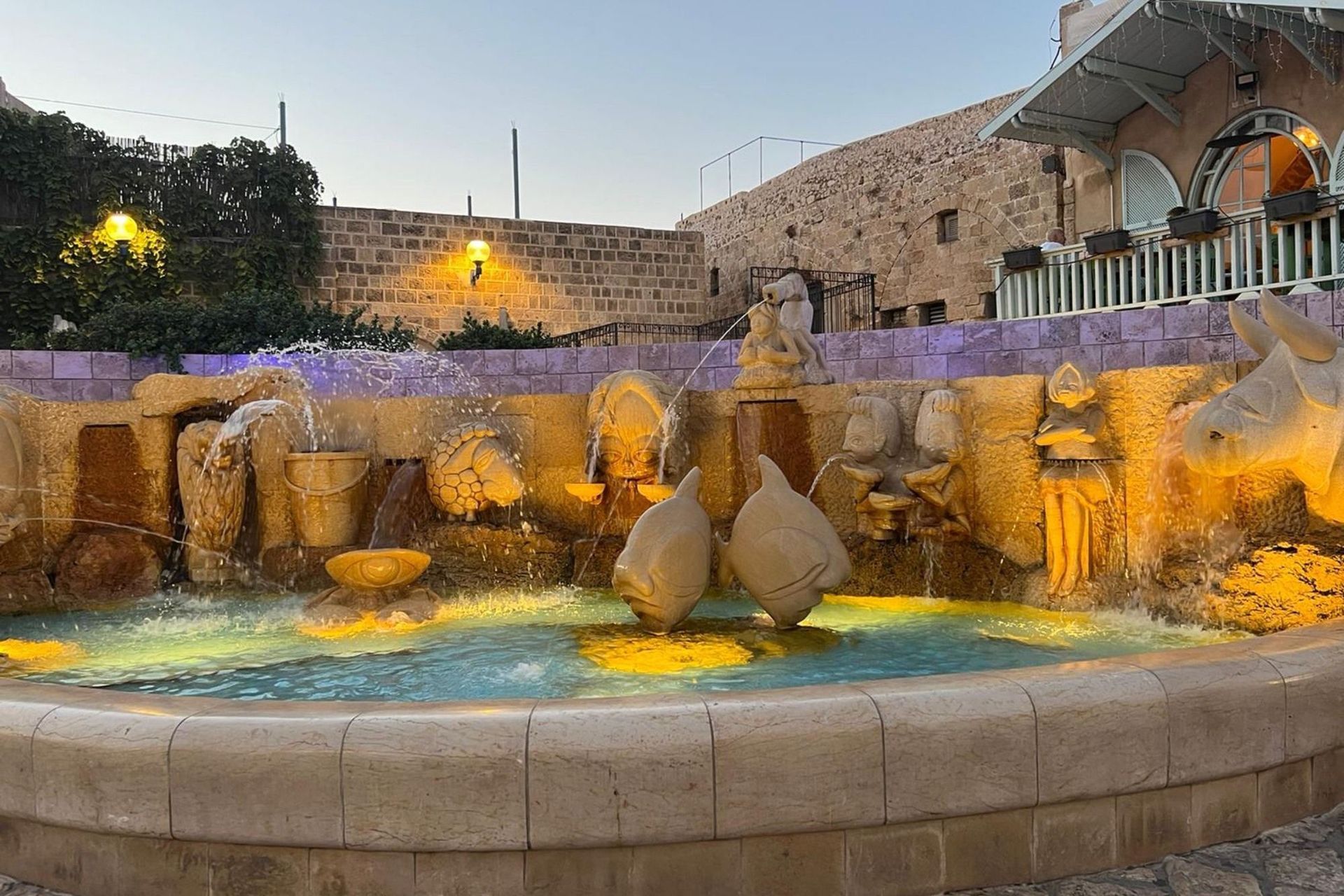 Fountain with statues and water jets in a stone courtyard, lit with warm and cool colors.
