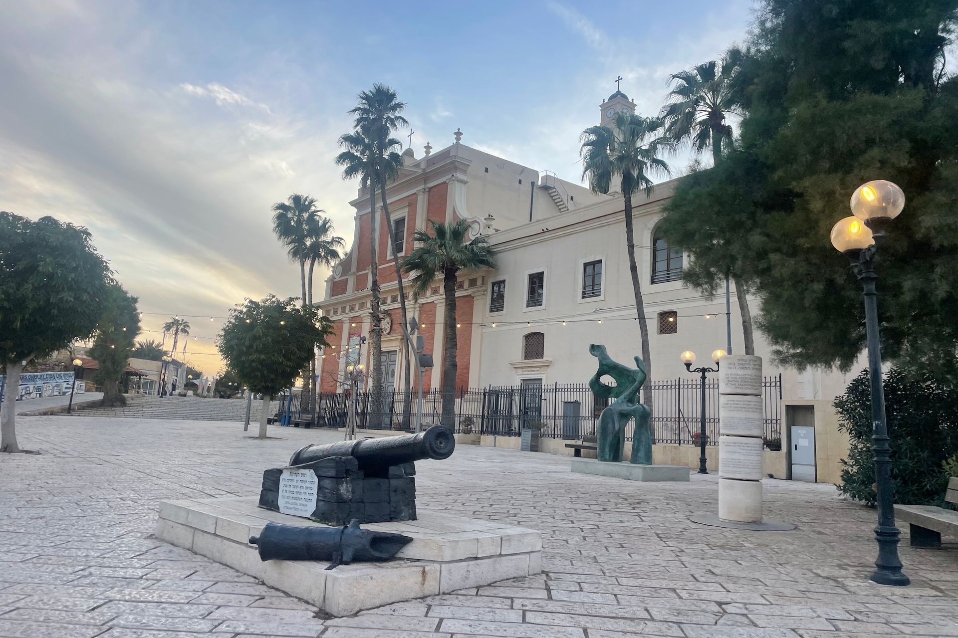 Historic square in Jaffa, Israel, with cannons, a church, sculptures, and palm trees against a cloudy sky.