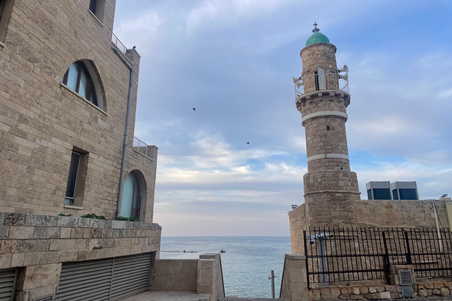 Stone buildings, including a minaret, line the coast under a cloudy sky.