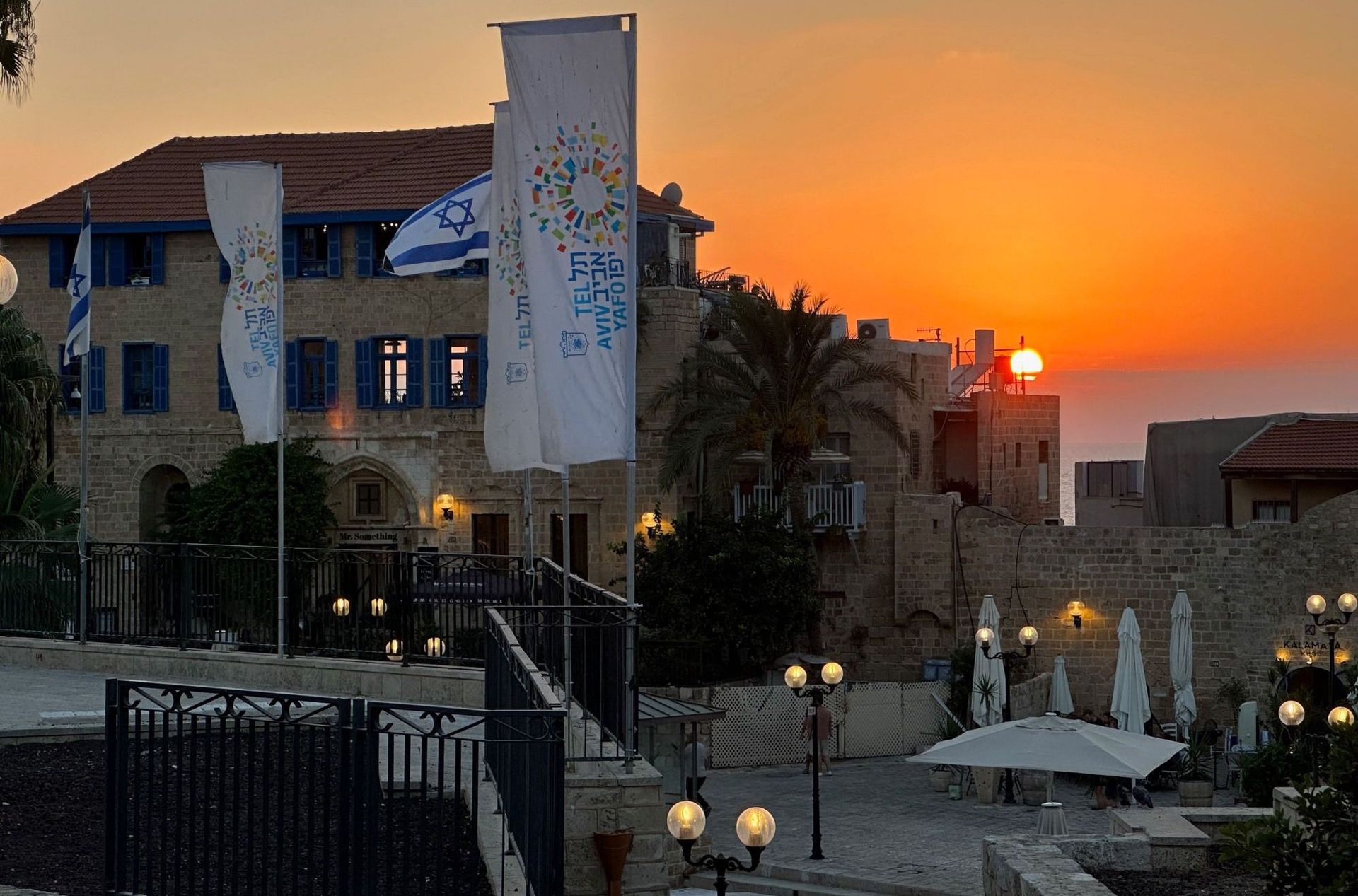 Sunset over a stone building with Israeli flags, blue shutters, and outdoor seating.