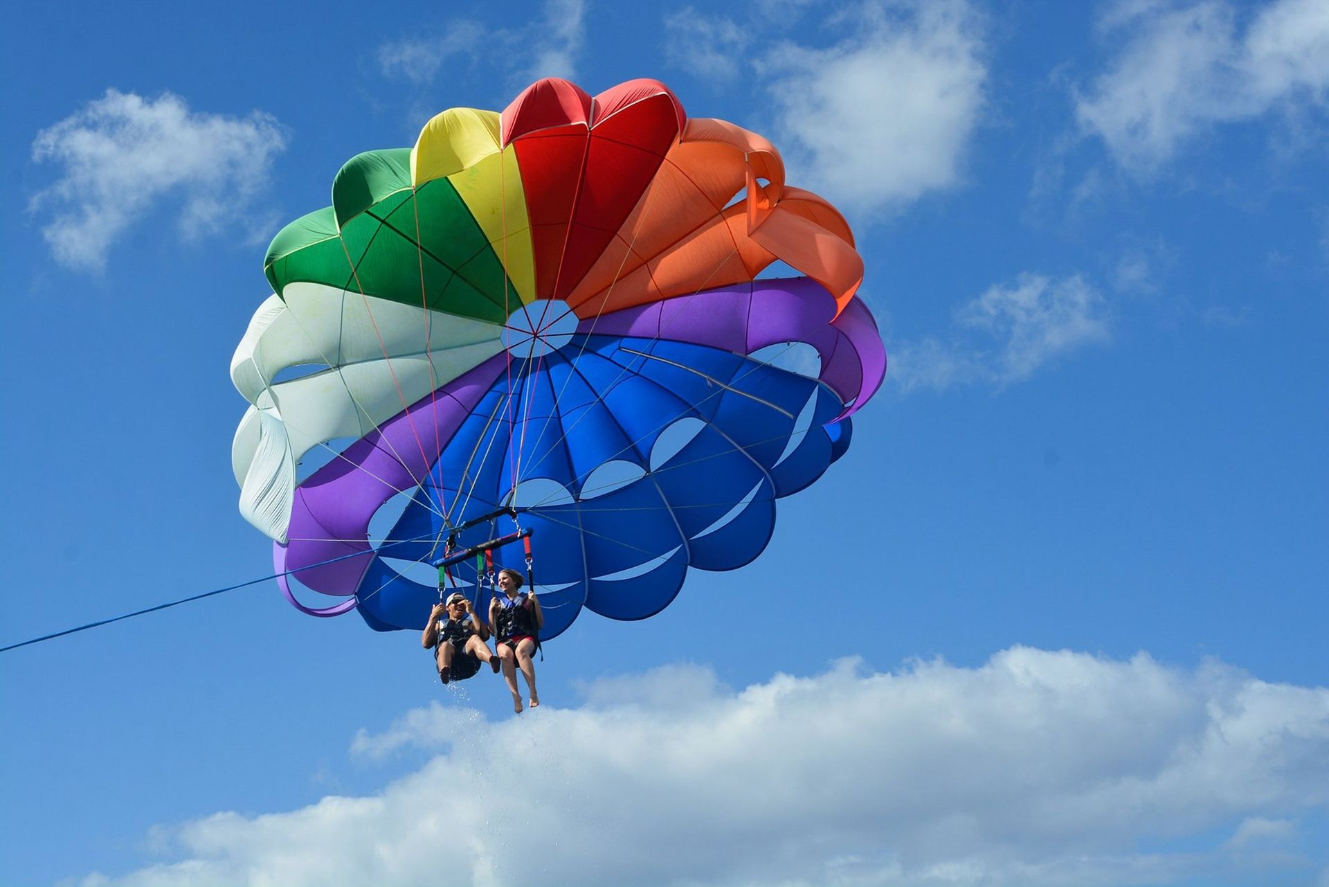 Two people parasailing under a colorful rainbow parachute against a blue sky with clouds.