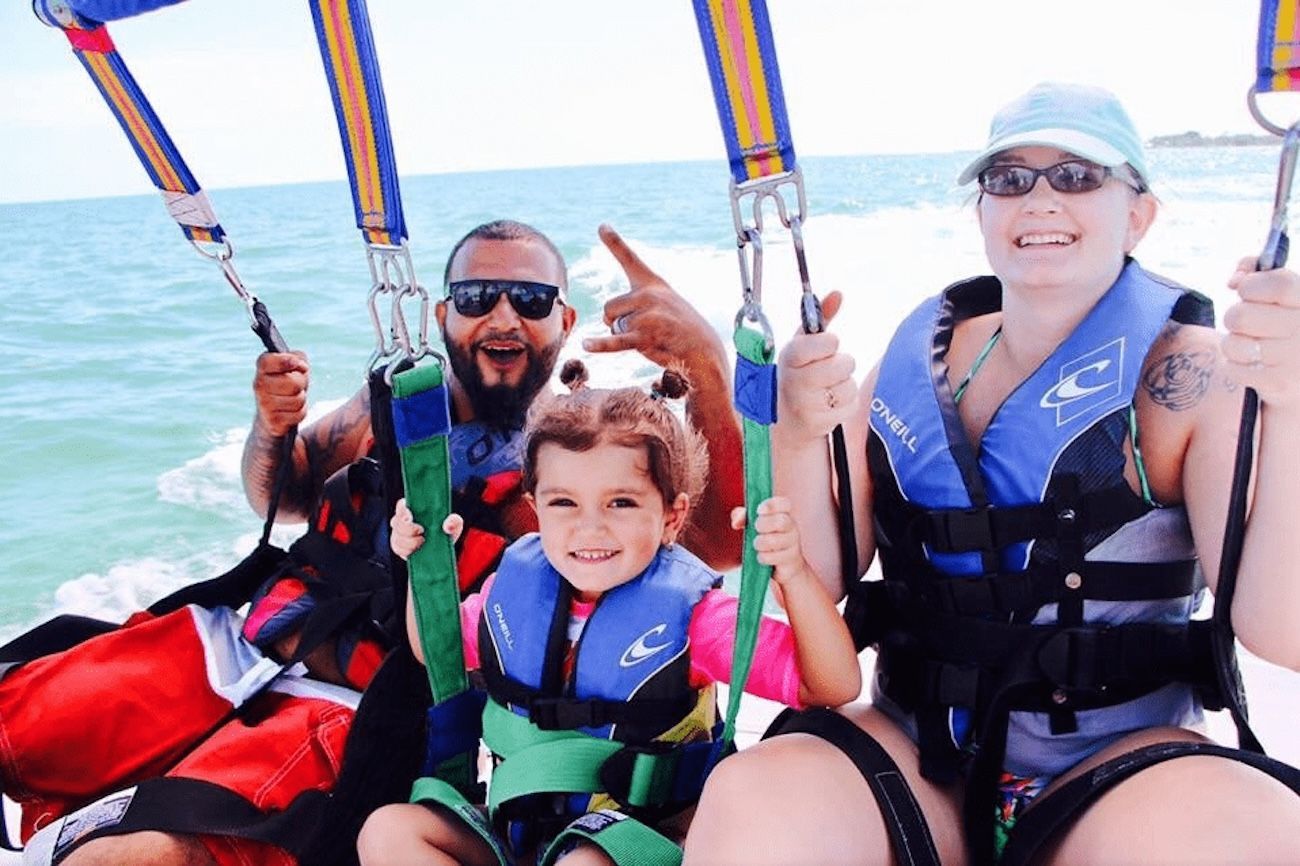 Family parasailing over ocean, smiling. Father gives hand gesture, wearing sunglasses.