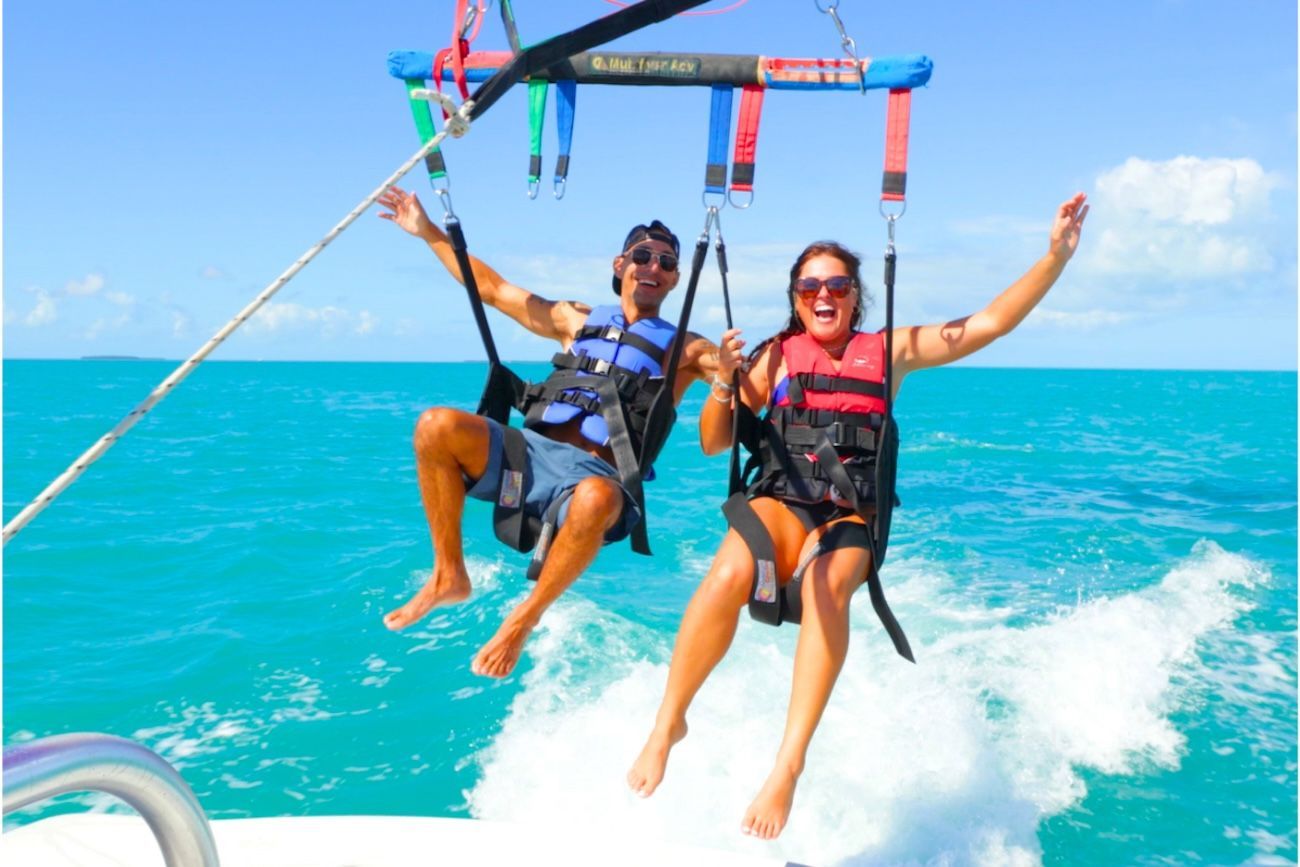 Two people parasailing over turquoise water, arms raised, smiling.