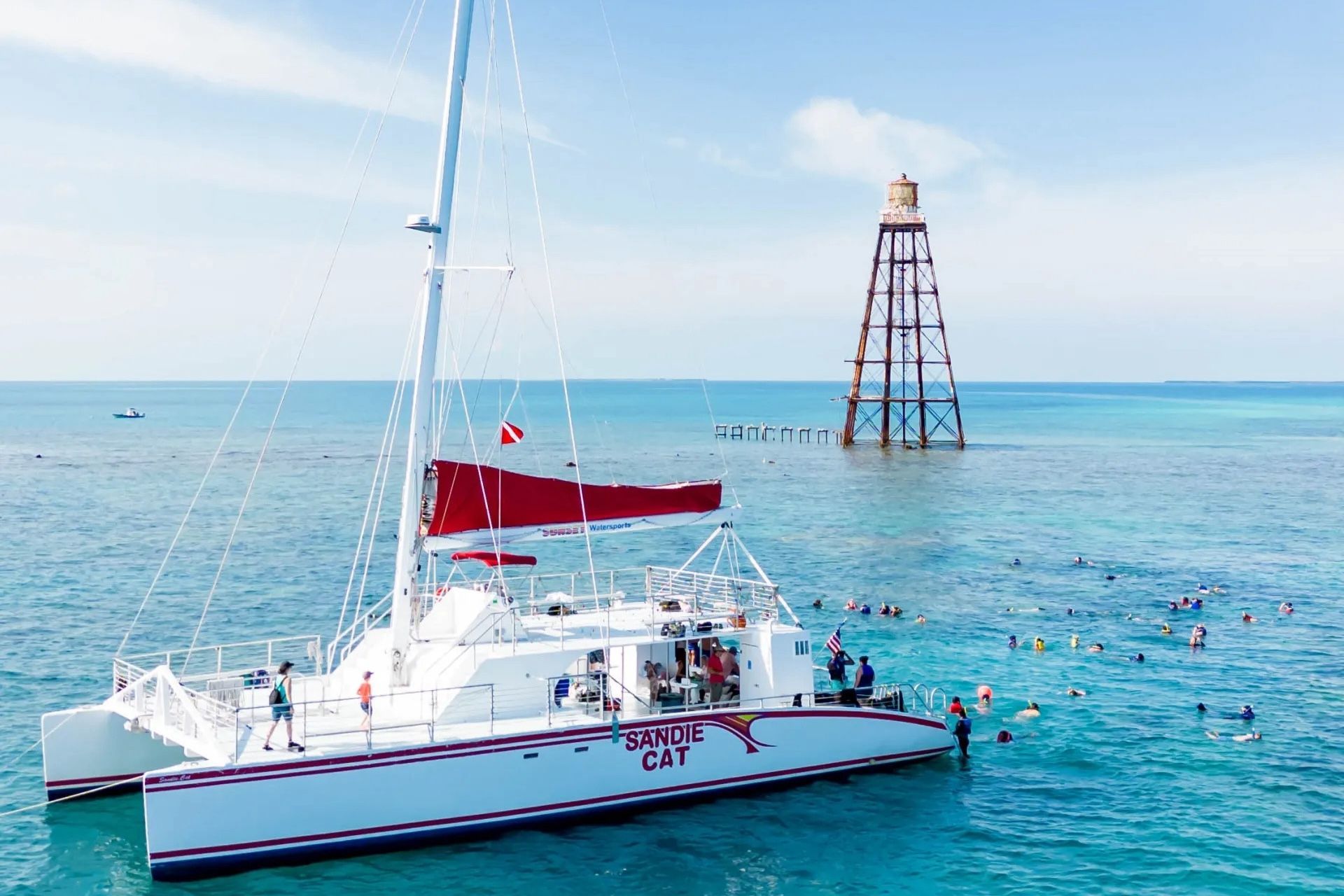 White catamaran sailboat near a structure in the ocean, snorkelers in the water, blue sky.